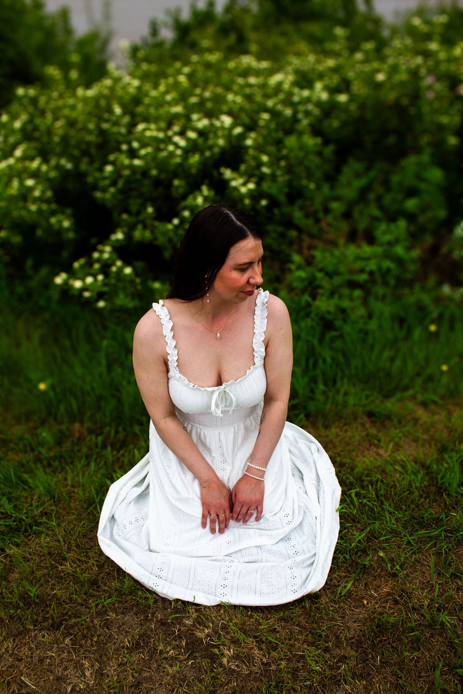 Woman in white dress kneeling on grass surrounded by lush green flowering bushes in a garden.