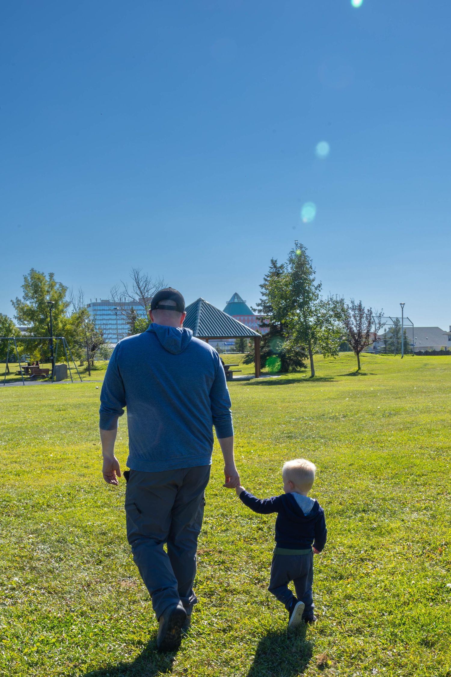 A father and young child walk hand-in-hand across a green park lawn under a bright blue sky.