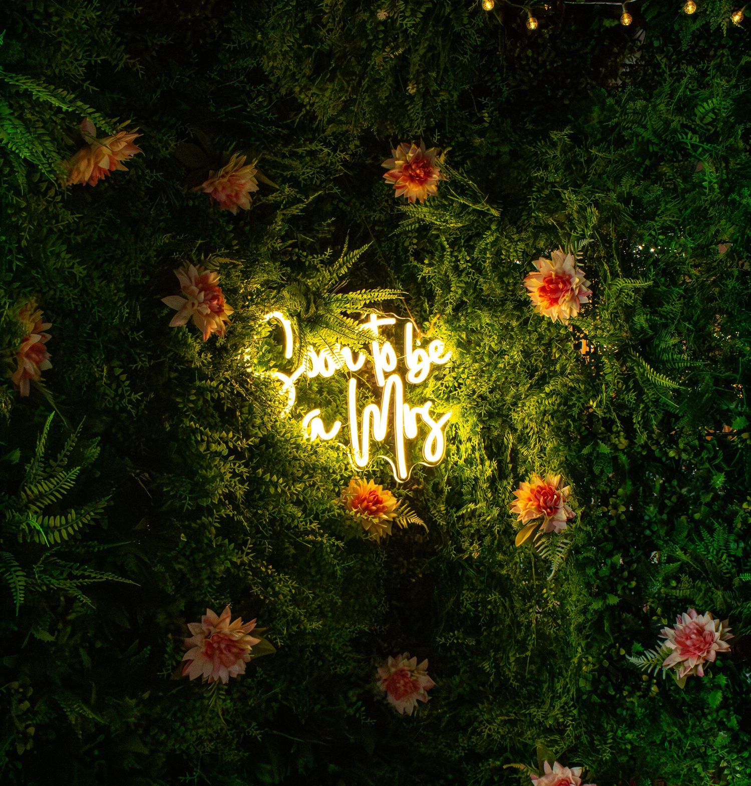 Glowing neon sign reading Soon to be a Mrs surrounded by lush greenery and pink flowers.