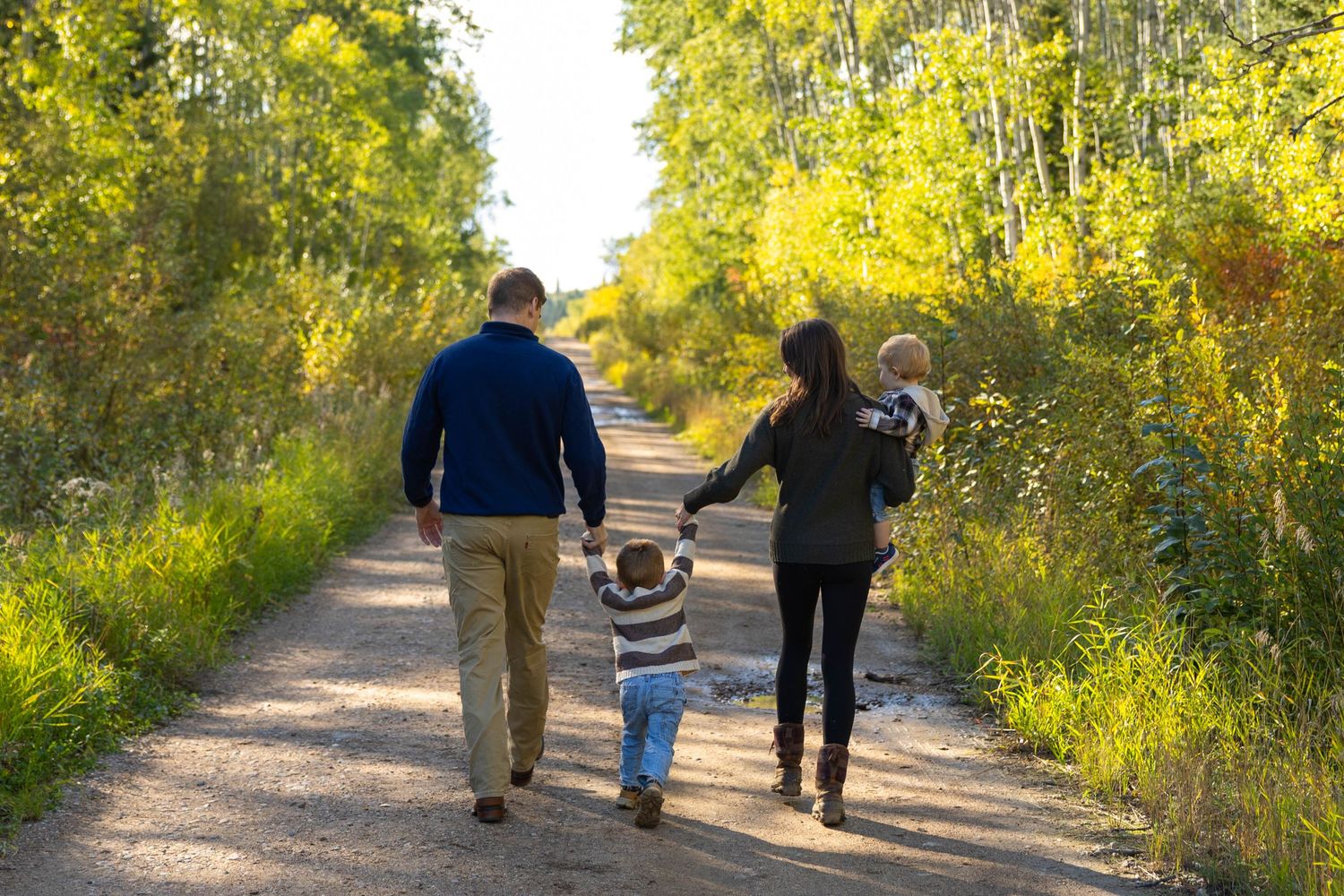 Family of four walking hand-in-hand down a scenic autumn trail surrounded by vibrant green and yellow trees.
