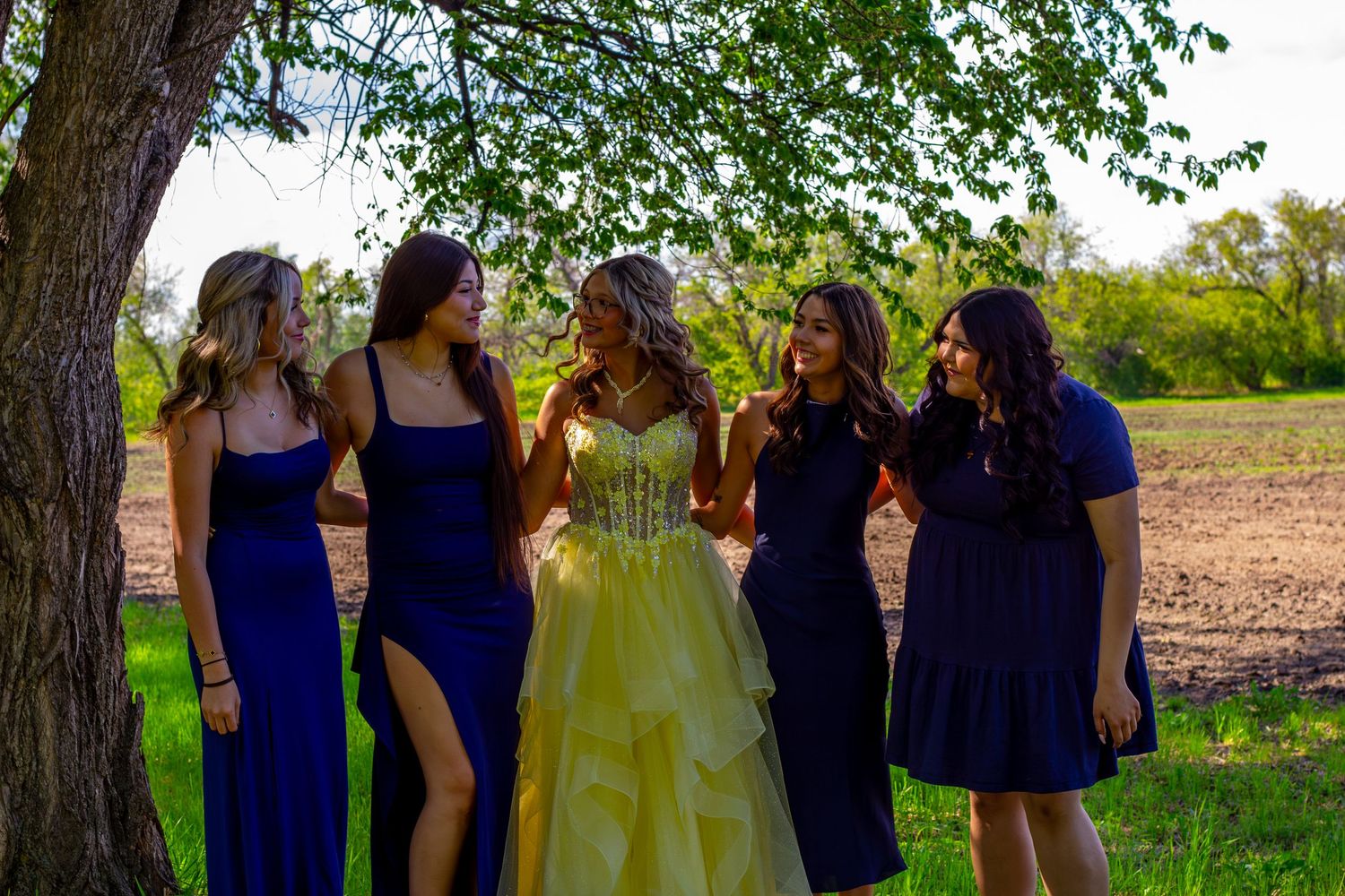Bride in yellow ball gown smiles with four bridesmaids in navy dresses beneath a leafy tree.