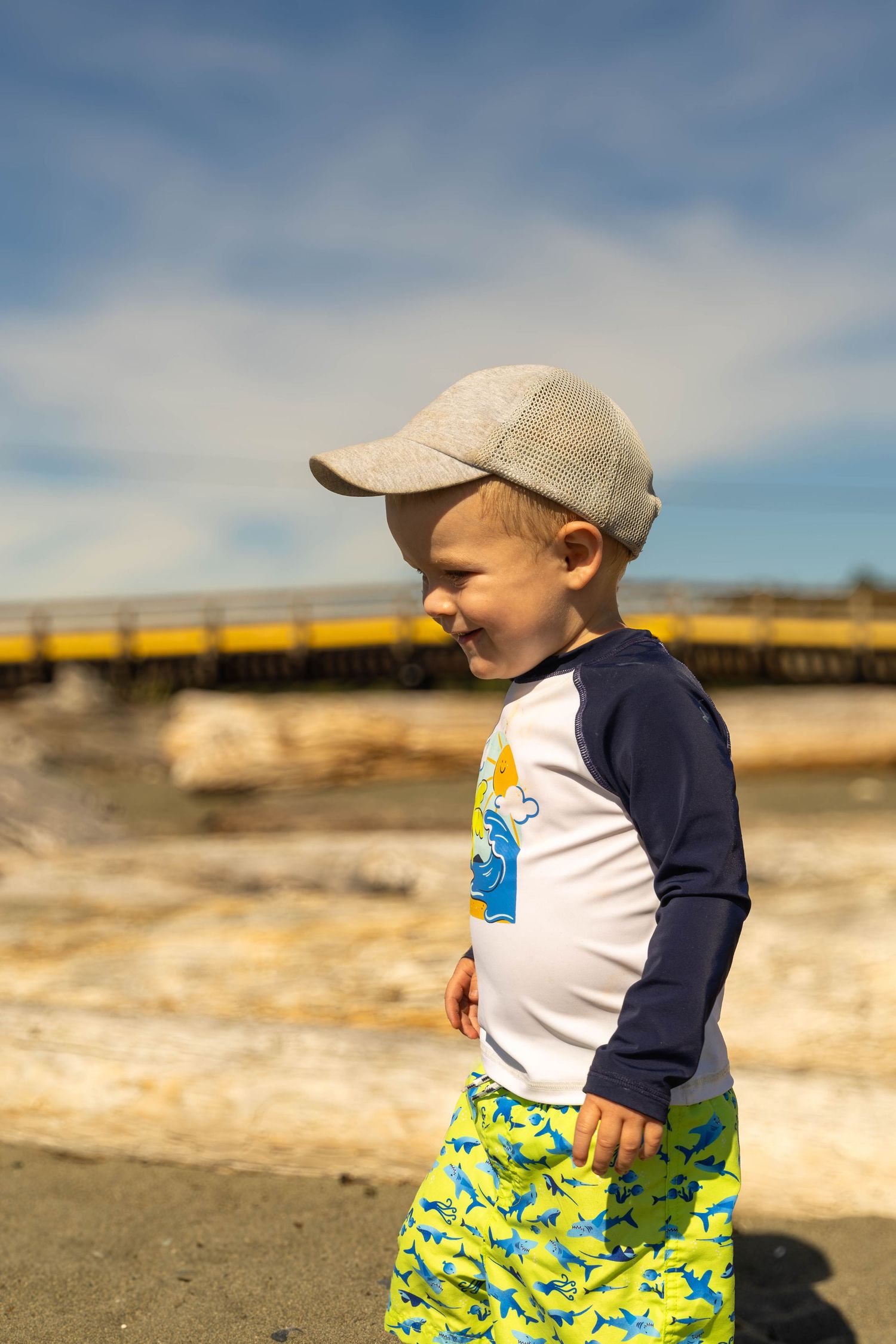 Happy toddler boy in cap and shark shorts smiling at sunny beach with bridge.