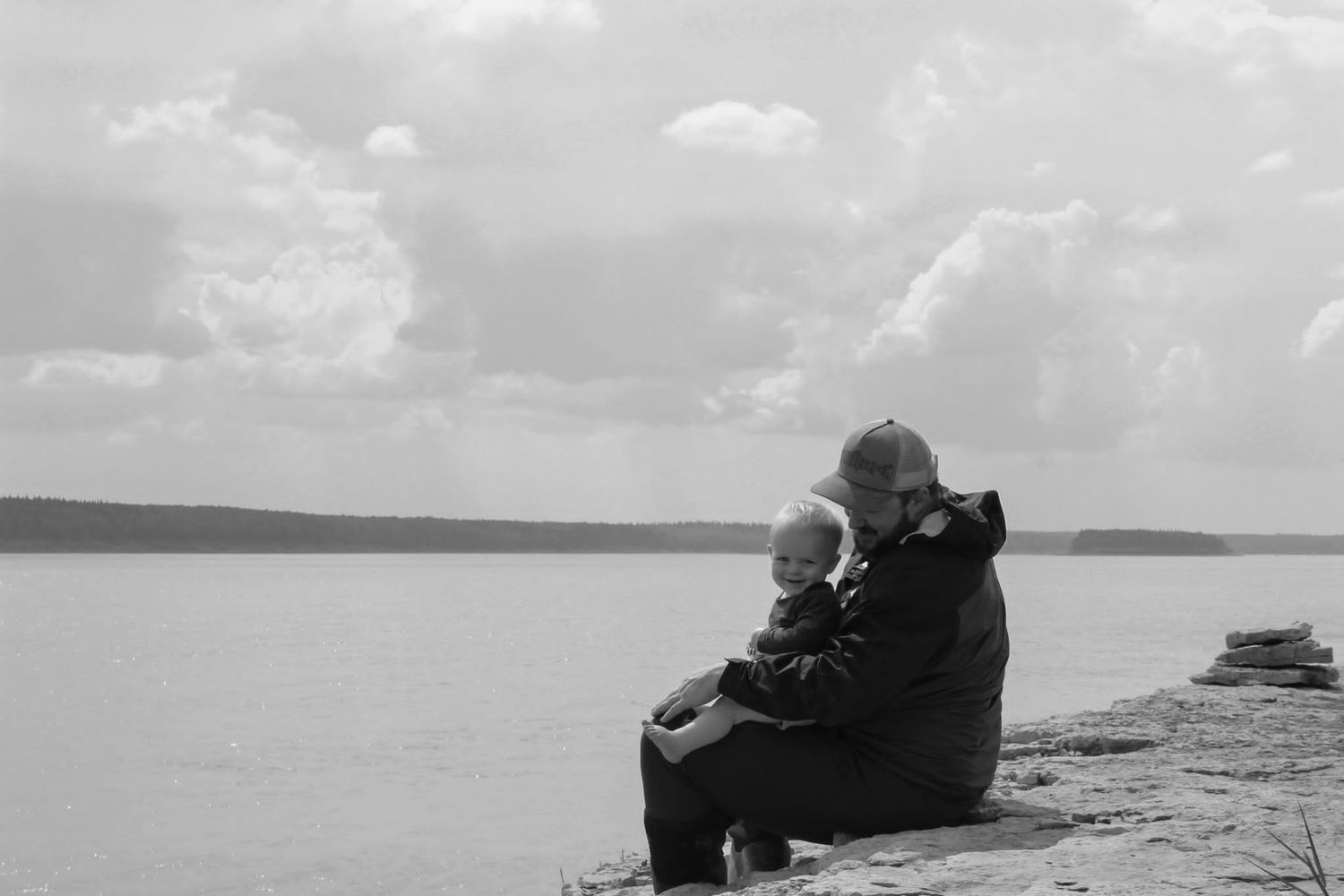 Black and white photo of adult and child sitting together on rocky lakeside under cloudy sky.