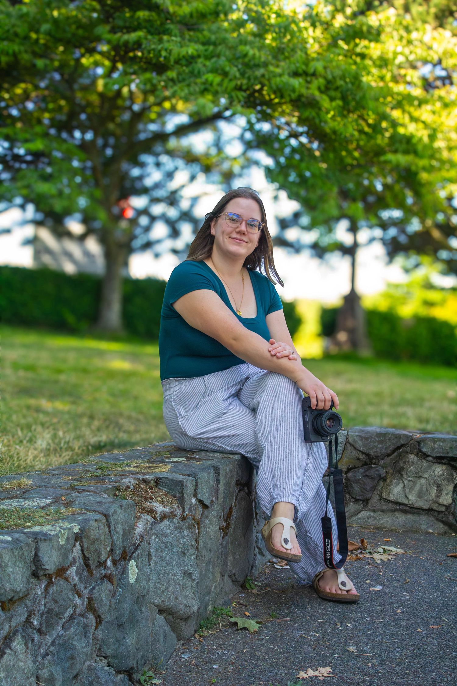 Young woman wearing glasses sits relaxed on a stone wall holding a camera in a green park.