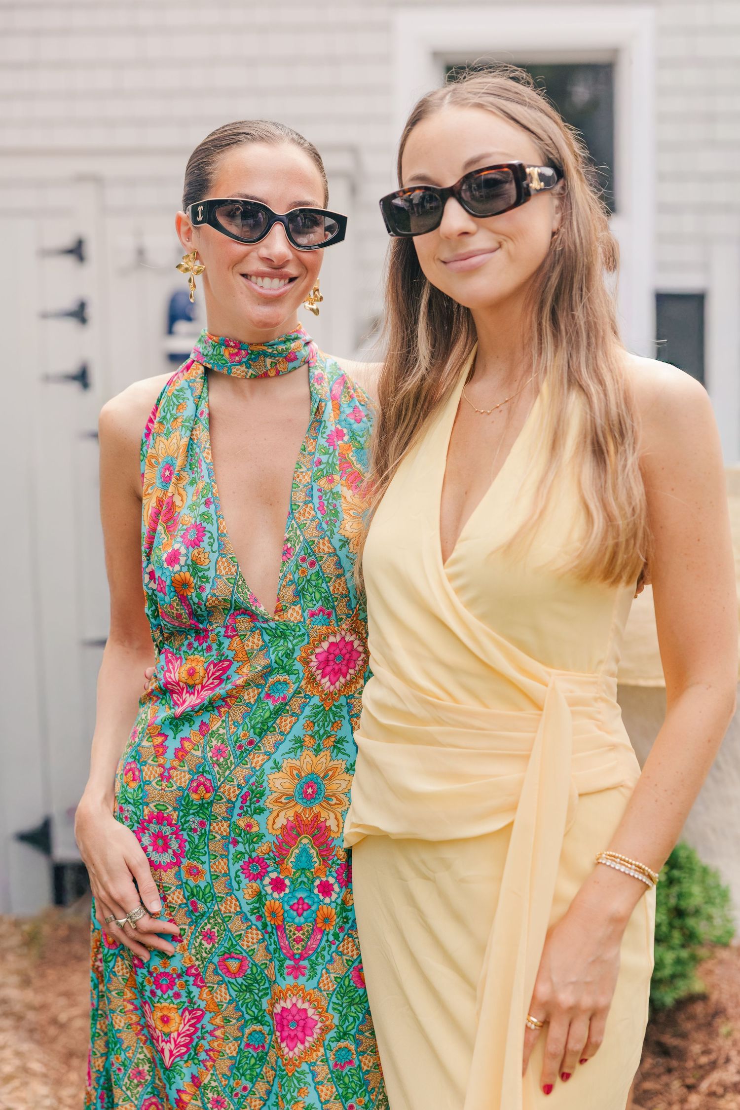 Two women wearing sunglasses pose outdoors, one in a colorful floral halter dress and one in a yellow wrap dress.