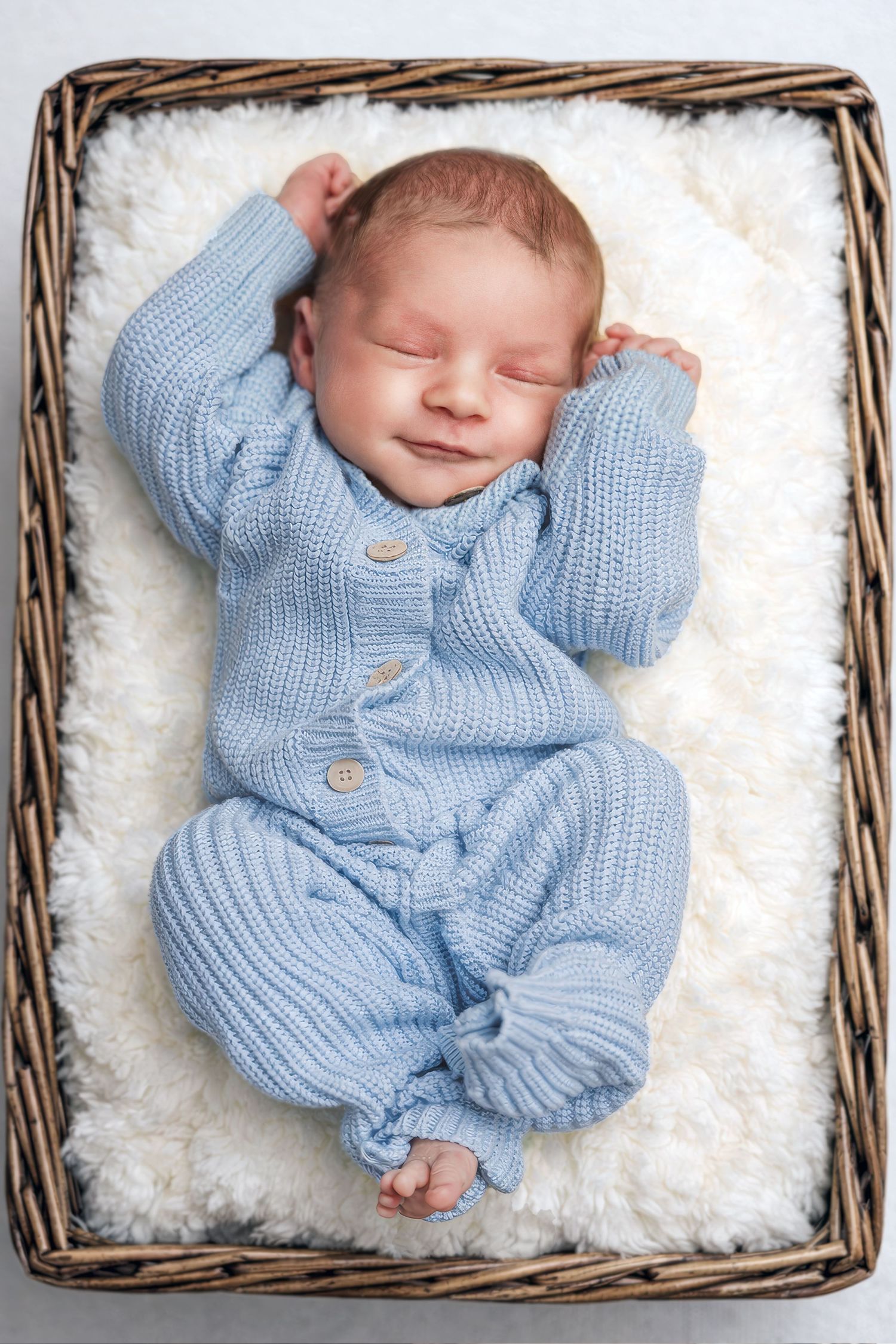 Smiling newborn baby in blue knit romper lying in wicker basket on white fluffy blanket.