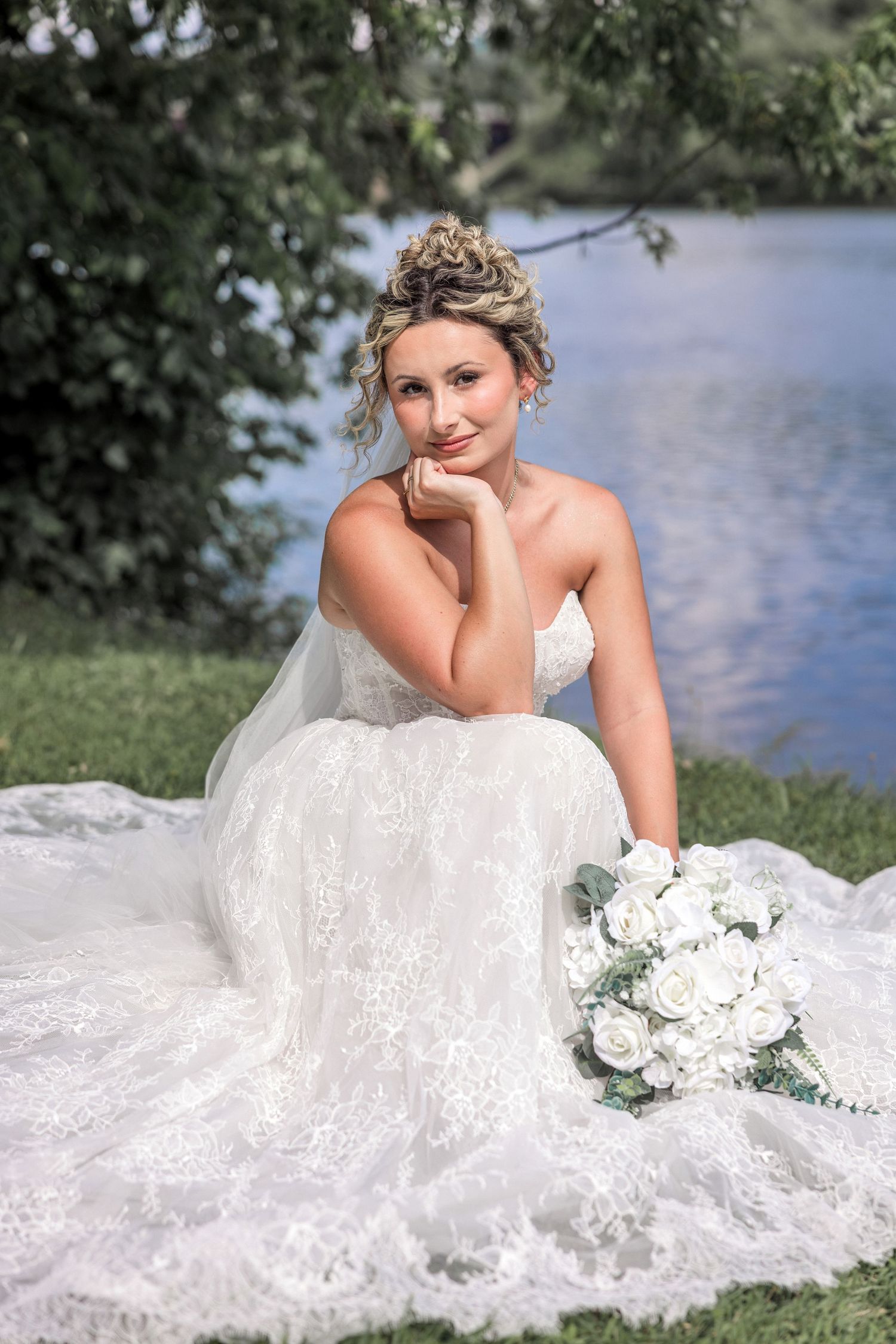 Bride in white lace gown sits by a lake holding a bouquet of white roses, smiling with hand resting on chin.