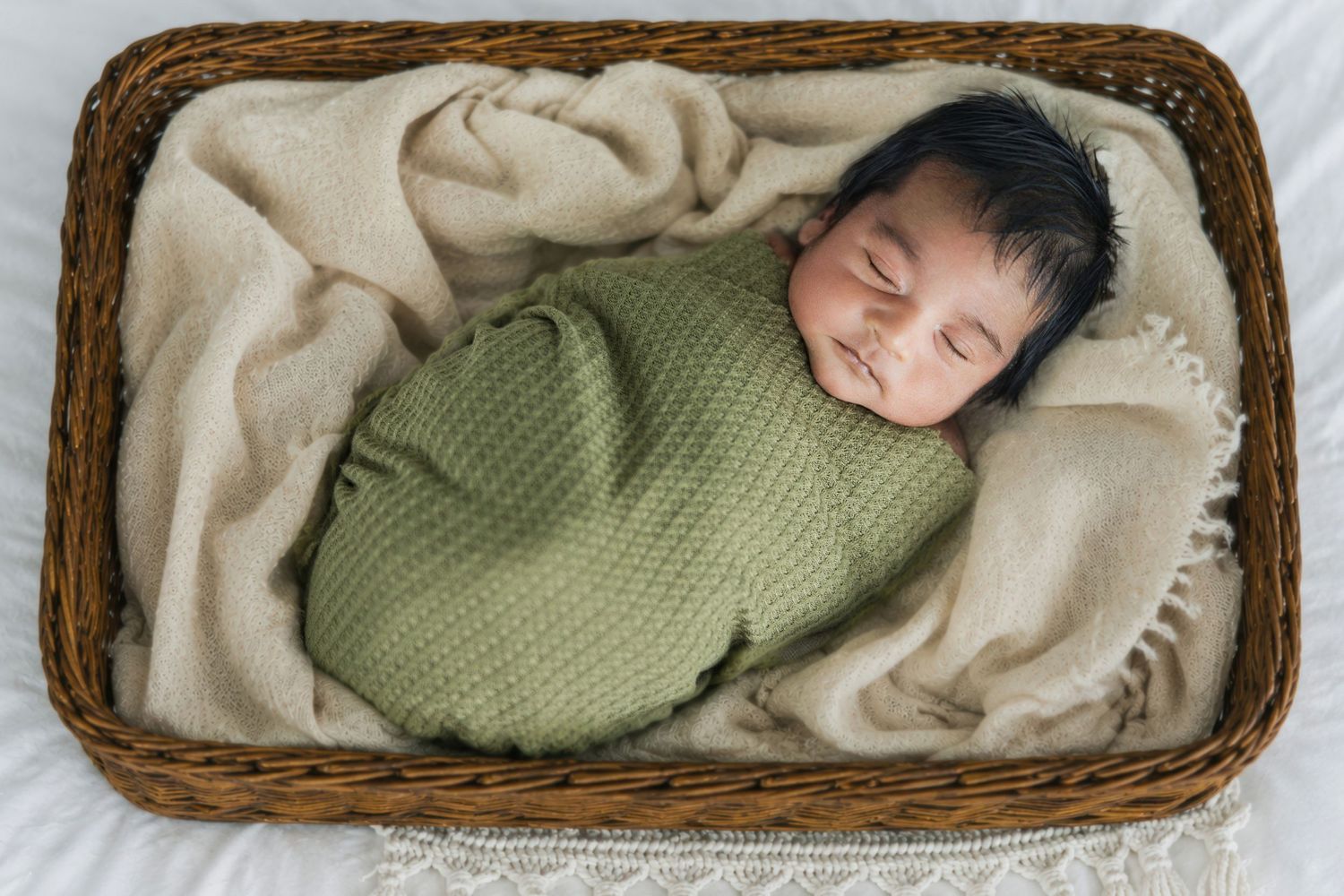 Peaceful newborn baby swaddled in green wrap sleeping in a wicker basket with beige blanket.