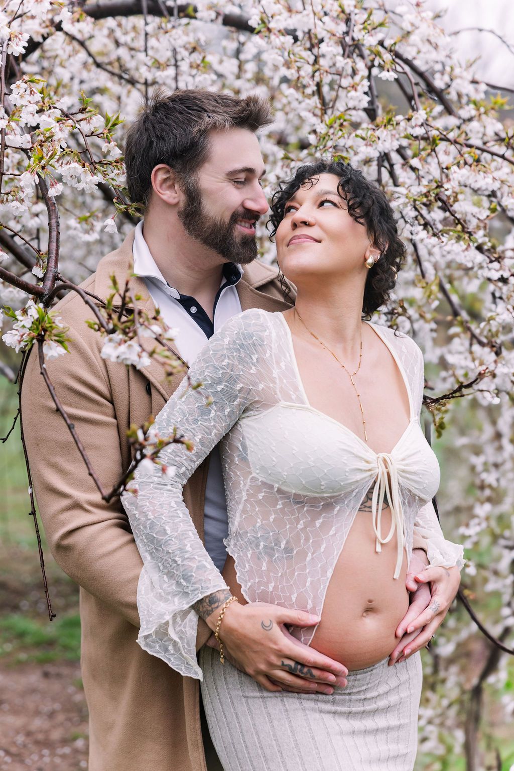 Couple embracing under cherry blossom trees, man holds pregnant woman's baby bump in spring garden.