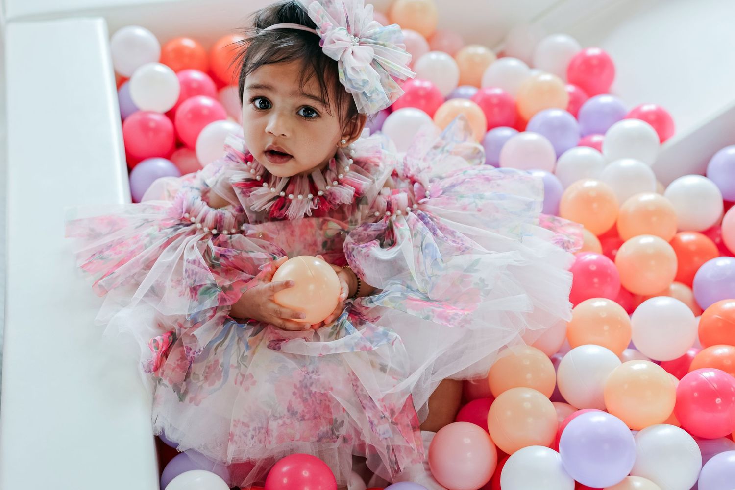Adorable toddler girl in floral tulle dress sitting in colorful ball pit at birthday party.
