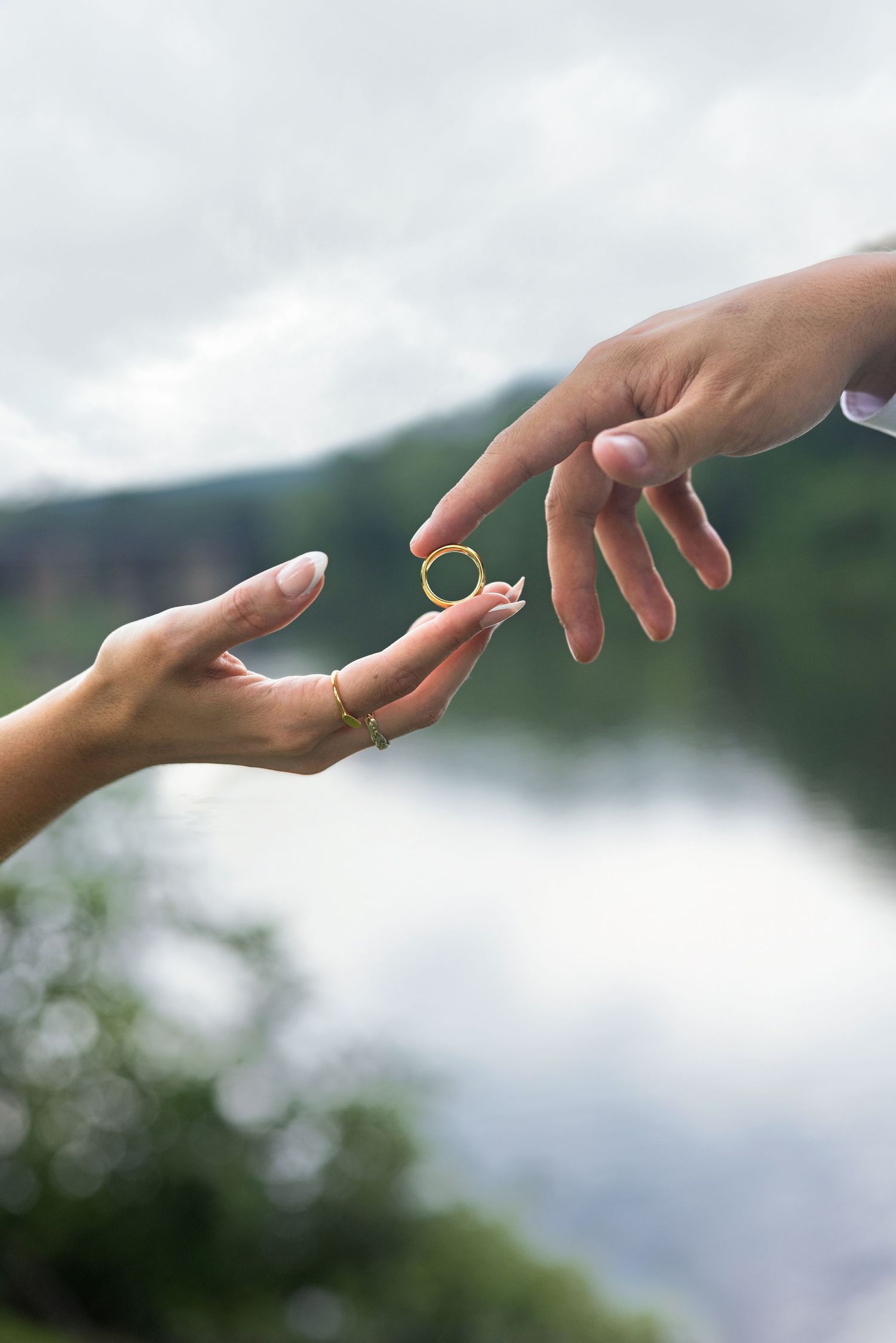 Two hands exchanging a gold wedding ring outdoors with a scenic lake and green mountains in the background.