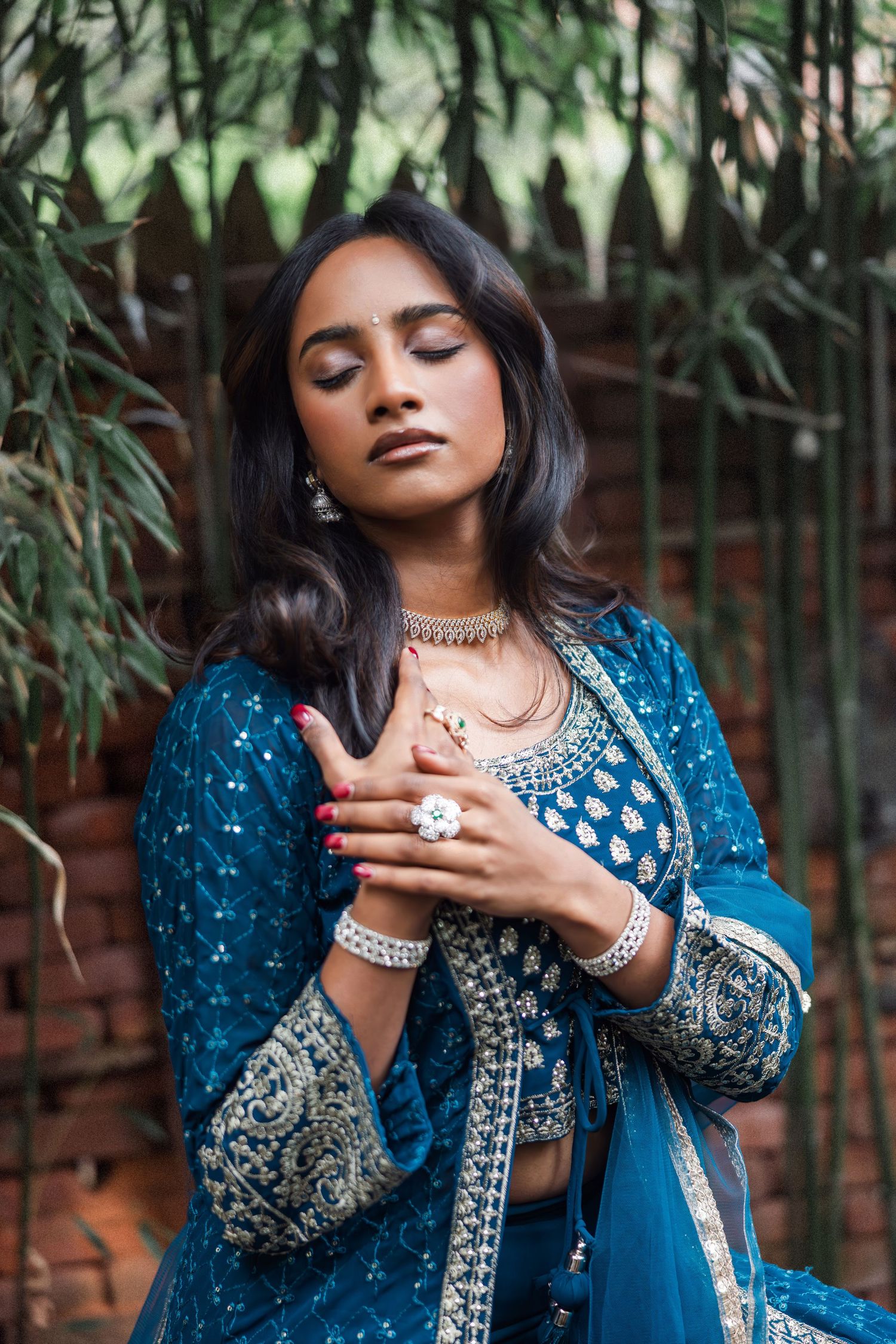 Woman in teal embroidered Indian lehenga with silver jewelry, eyes closed, posing gracefully outdoors.