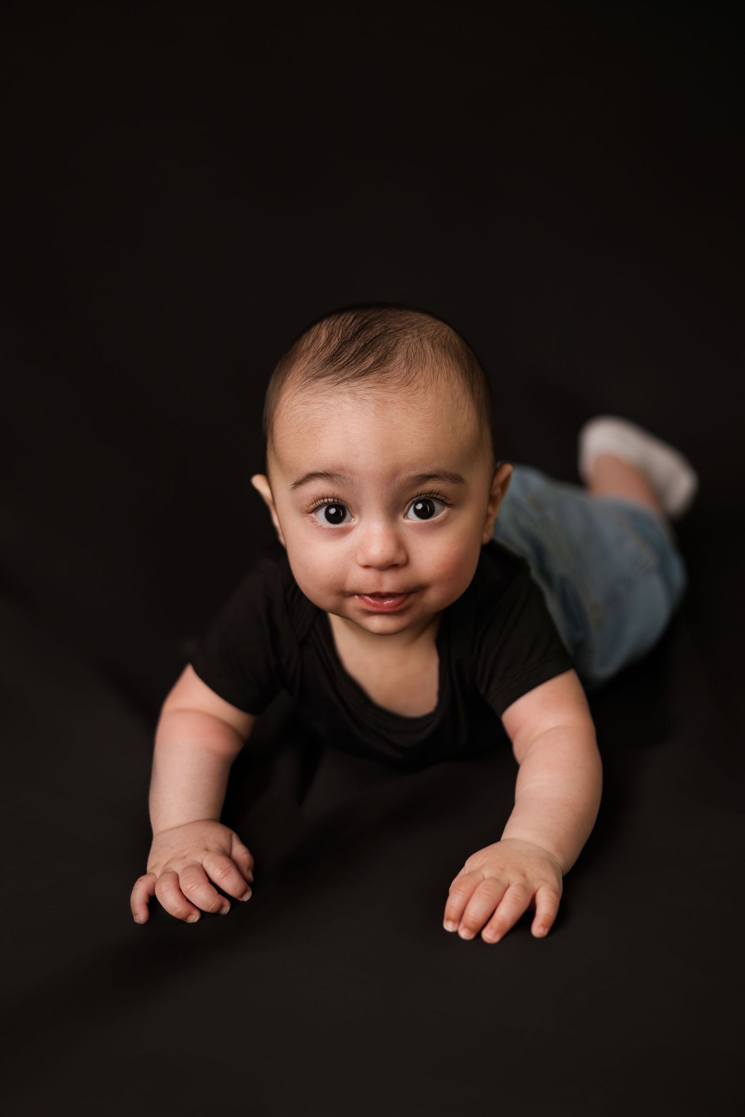 Adorable baby in black onesie and jeans crawling on dark floor, looking up at camera with big eyes.