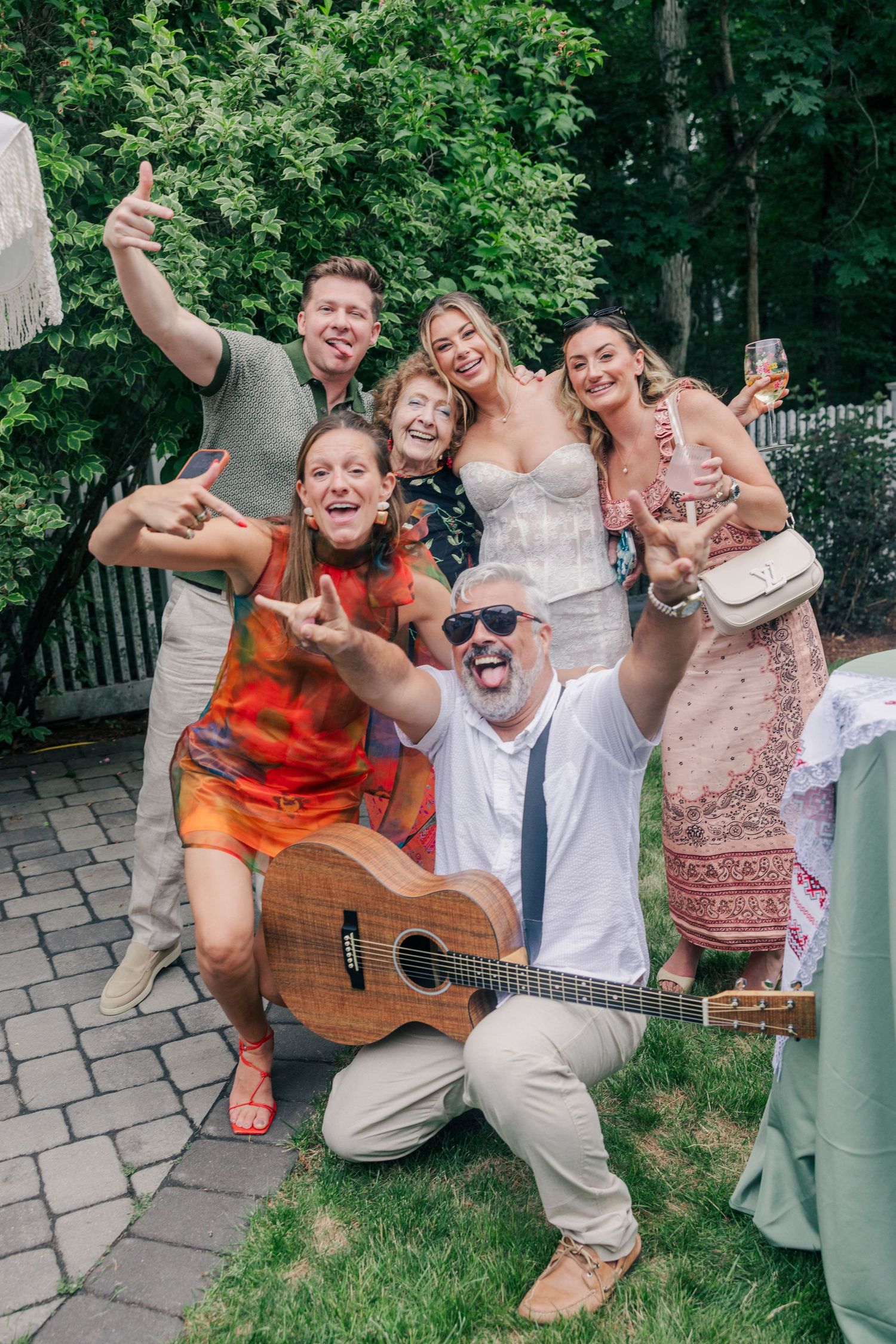 Group of six people posing joyfully at an outdoor wedding, one man holding an acoustic guitar on a garden patio.