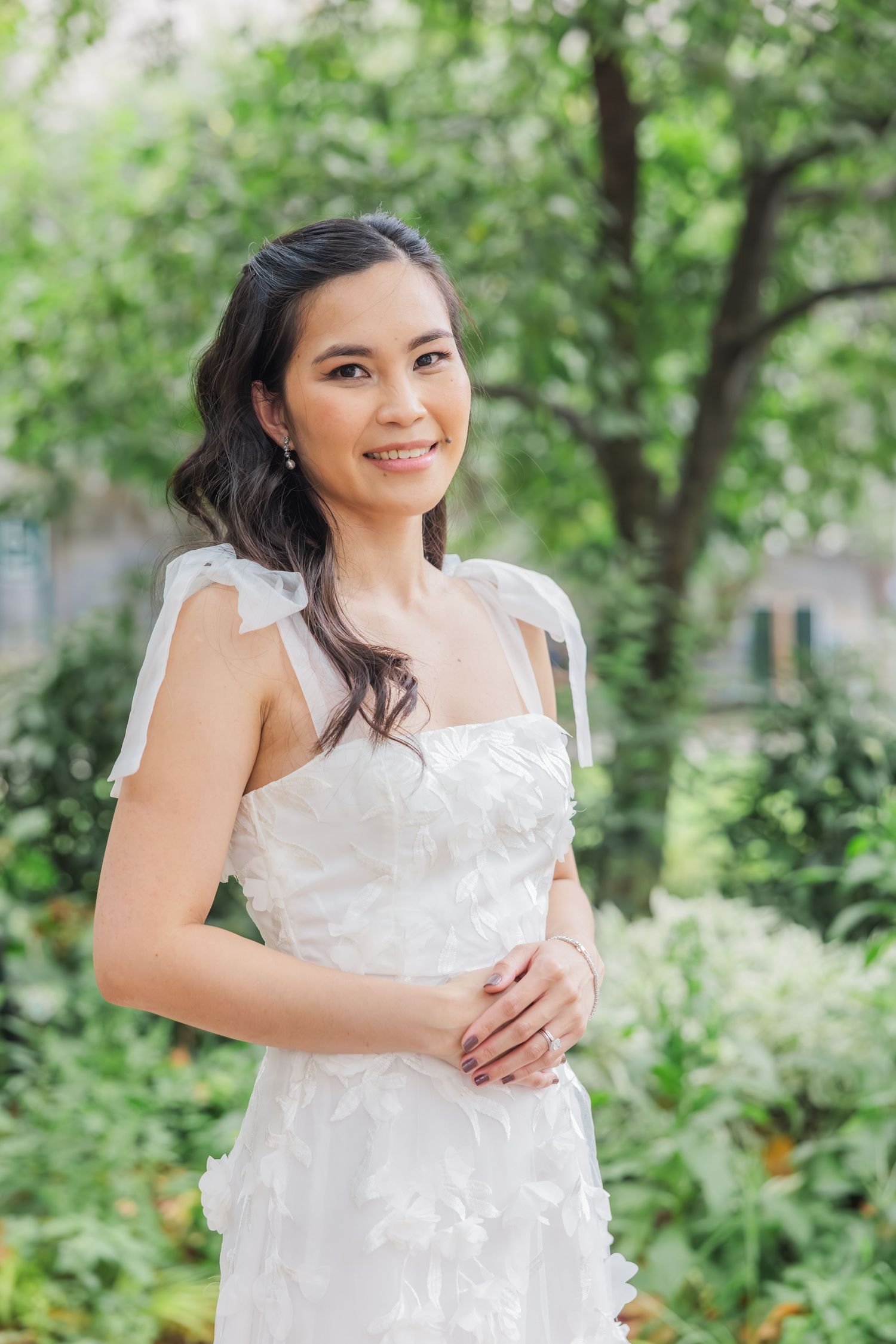 Smiling Asian bride in a white floral wedding dress with bow straps posing in a lush garden.