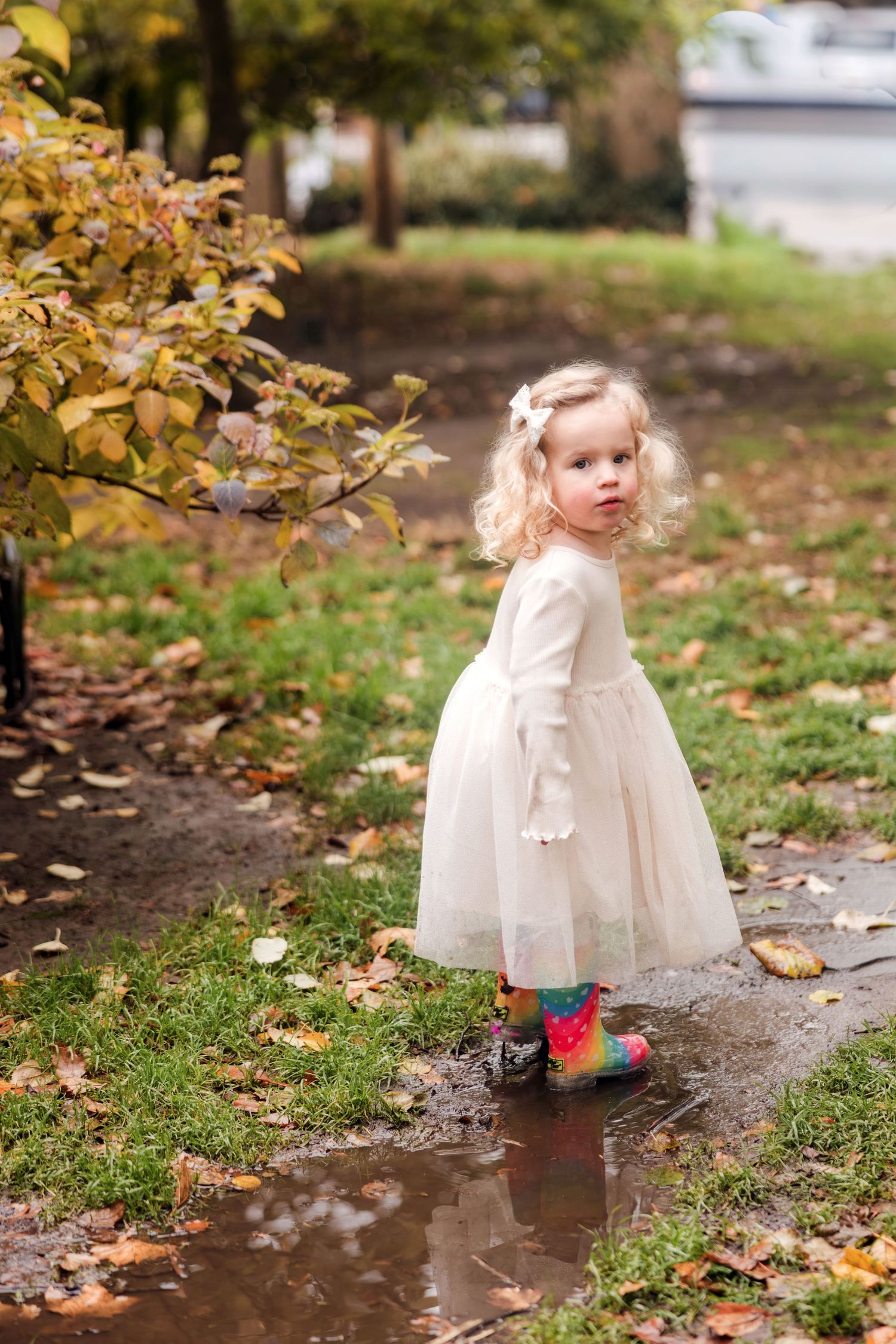 Curly-haired toddler in white dress and rainbow boots splashing in puddle during autumn day.