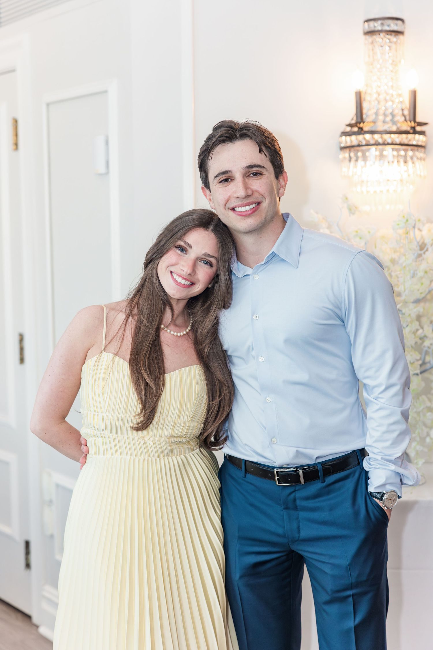Smiling couple posing together, woman in yellow pleated dress and man in blue shirt and pants, elegant interior.
