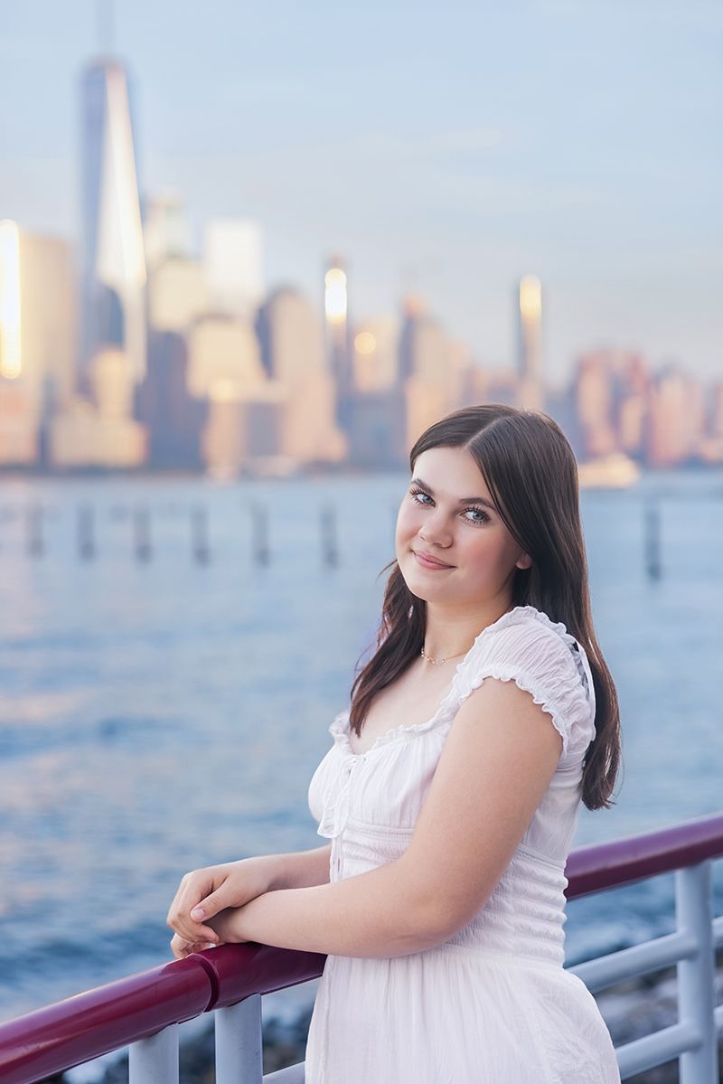 Young woman in white dress leaning on railing with NYC skyline and Hudson River in background at dusk.