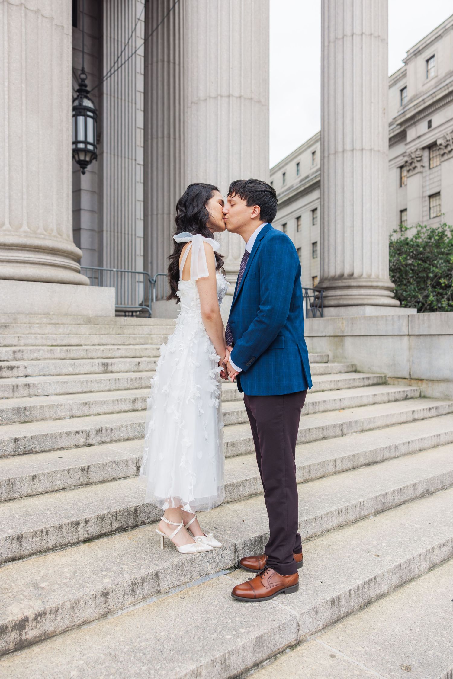 Bride and groom kissing on stone steps of a grand building with tall columns on their wedding day.