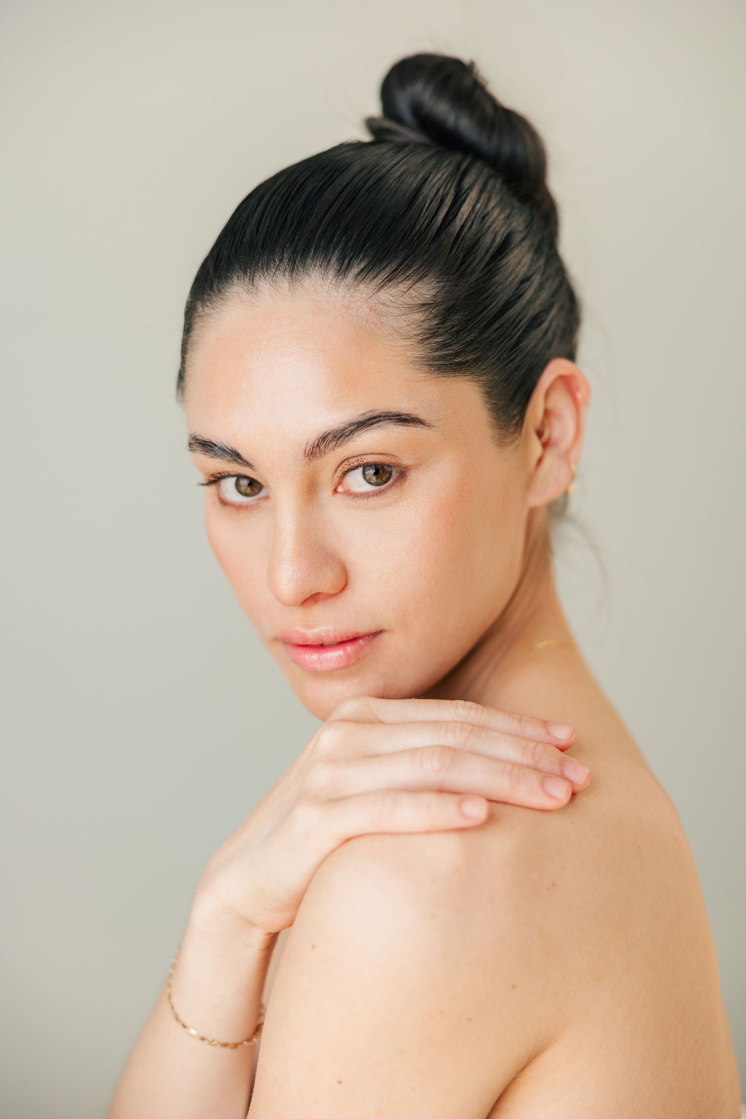 Woman with dark hair in bun touching shoulder, minimalist beauty portrait on gray background.