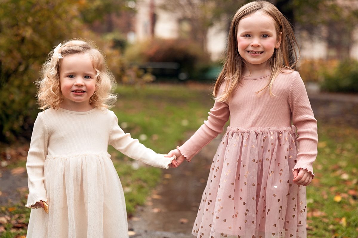 Two young sisters in pink dresses holding hands and smiling outdoors in autumn park.