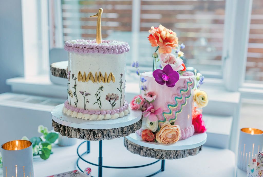Two decorated first birthday cakes with floral designs on rustic wood stands in bright room.