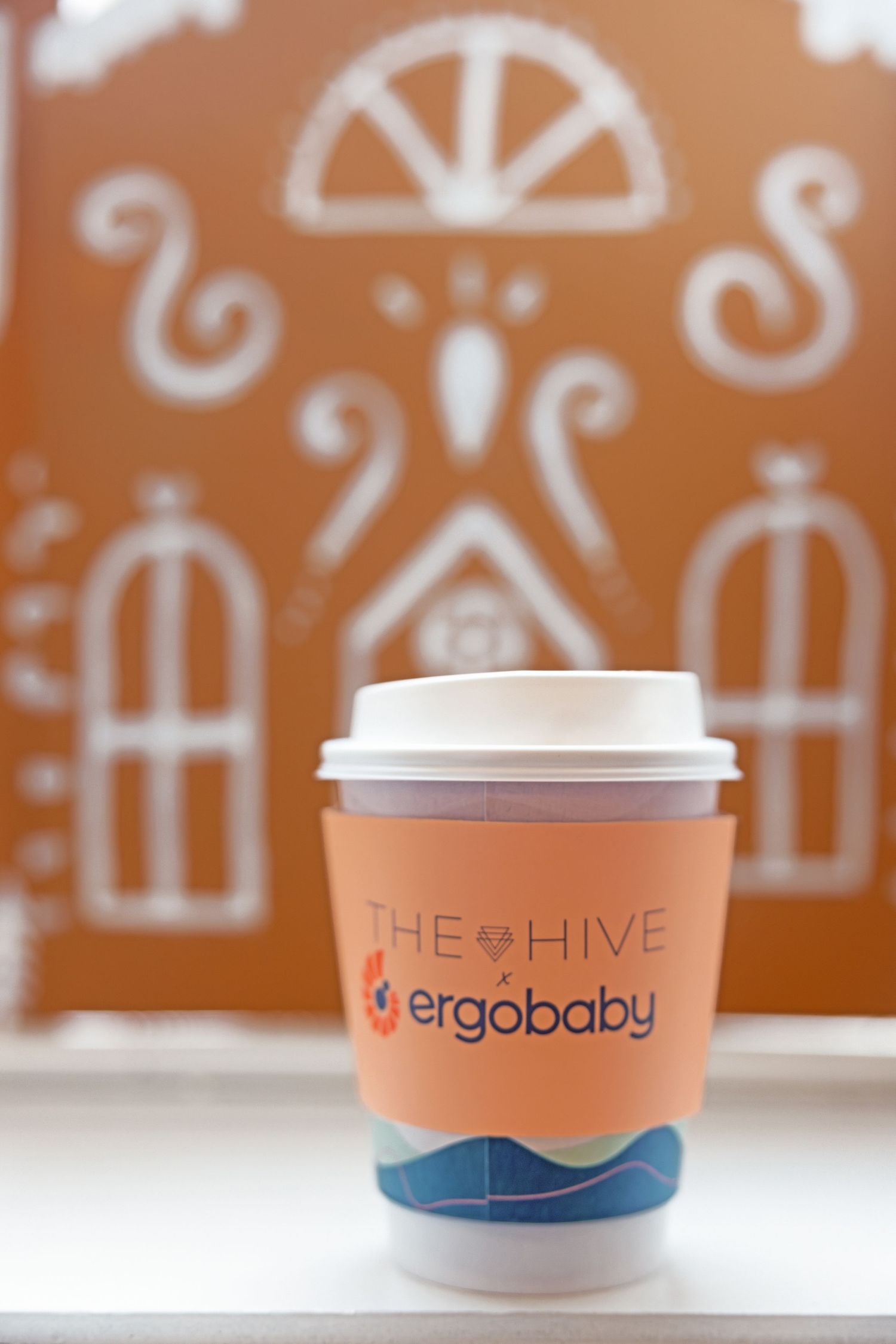 Ergobaby and The Hive branded orange coffee cup with white lid on a windowsill with decorative background.