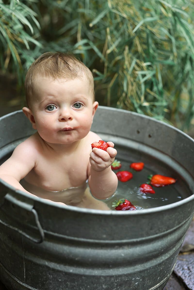Cute baby sitting in vintage galvanized tub outdoors holding a strawberry surrounded by floating berries.