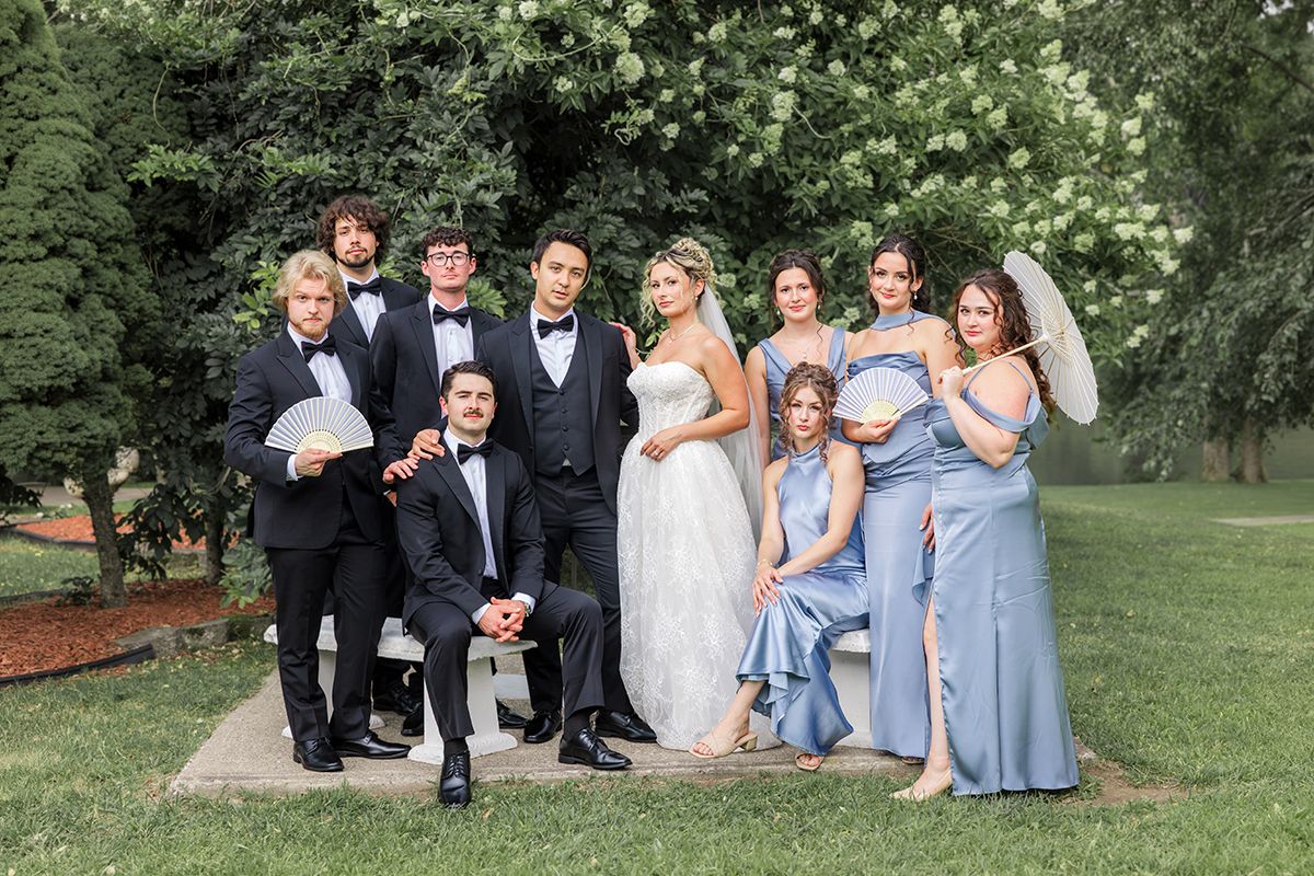 Wedding party poses outdoors in formal attire, bridesmaids in blue gowns flanking bride and groom in lush garden setting.
