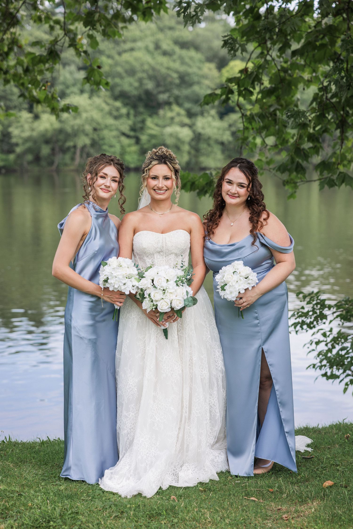 Bride in white lace gown flanked by two bridesmaids in blue satin dresses holding white bouquets by a lake.