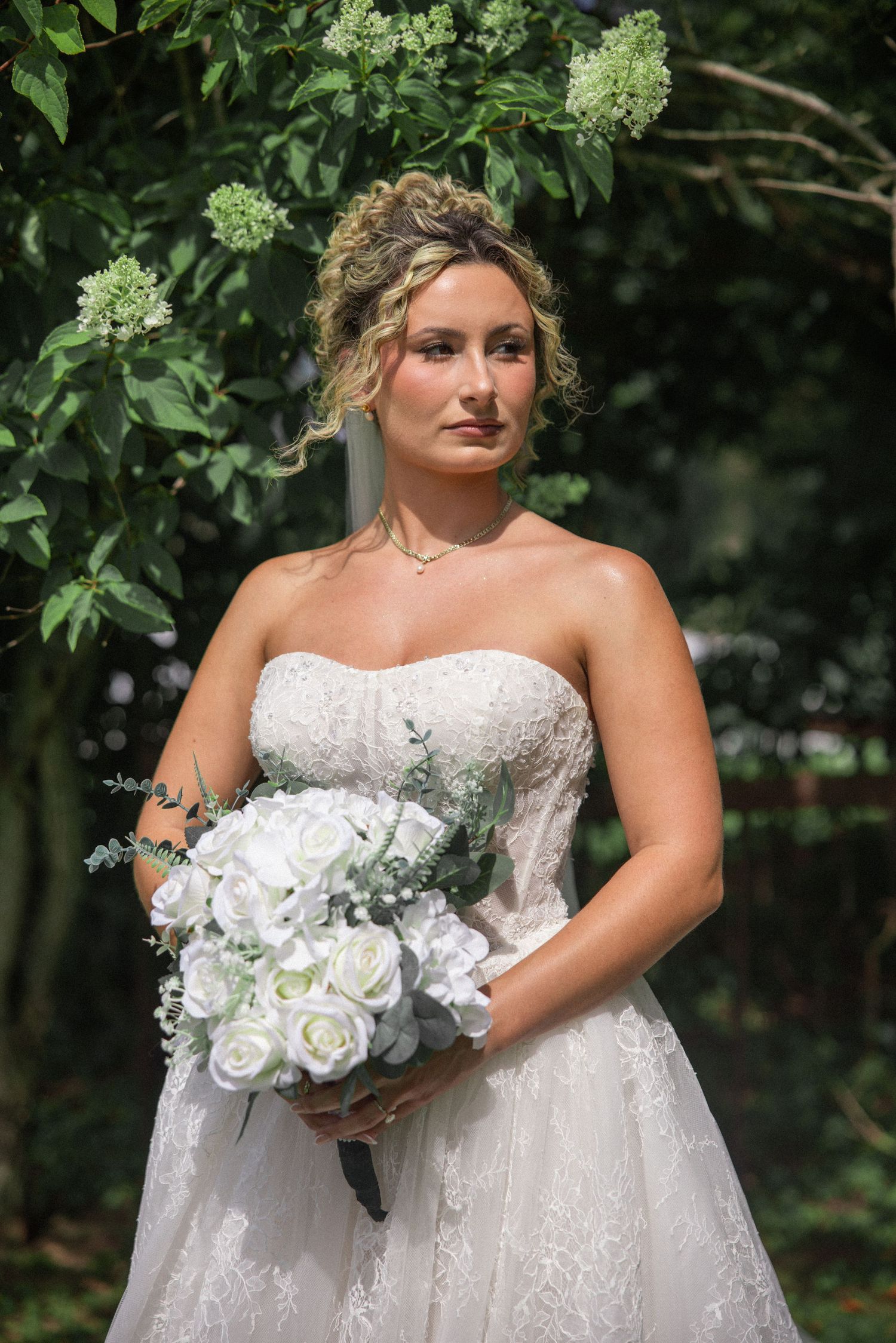 Bride in strapless lace wedding gown holding white rose bouquet, posing outdoors among lush green foliage.