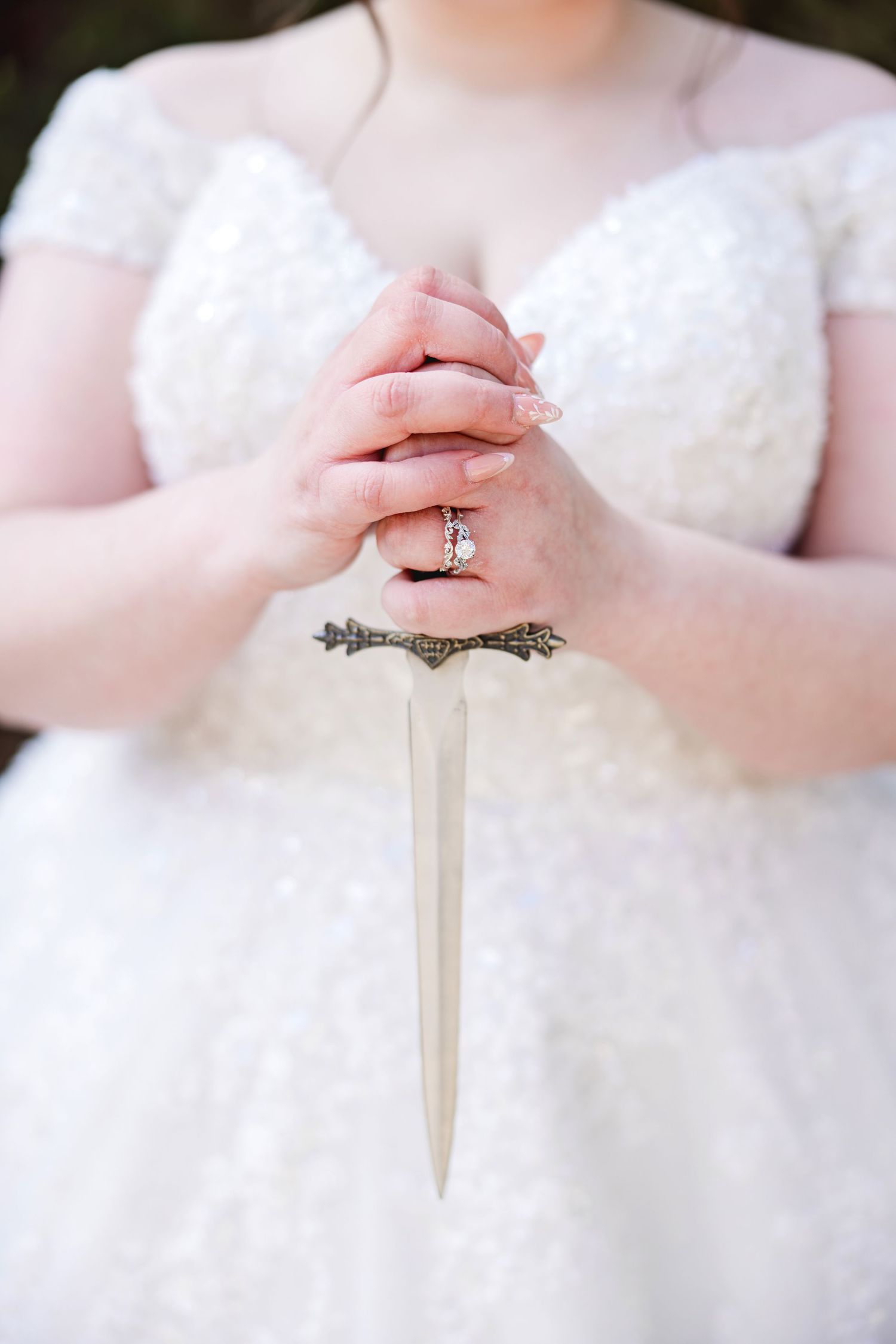 Bride in white lace gown holding an ornate dagger with clasped hands, wearing an engagement ring.
