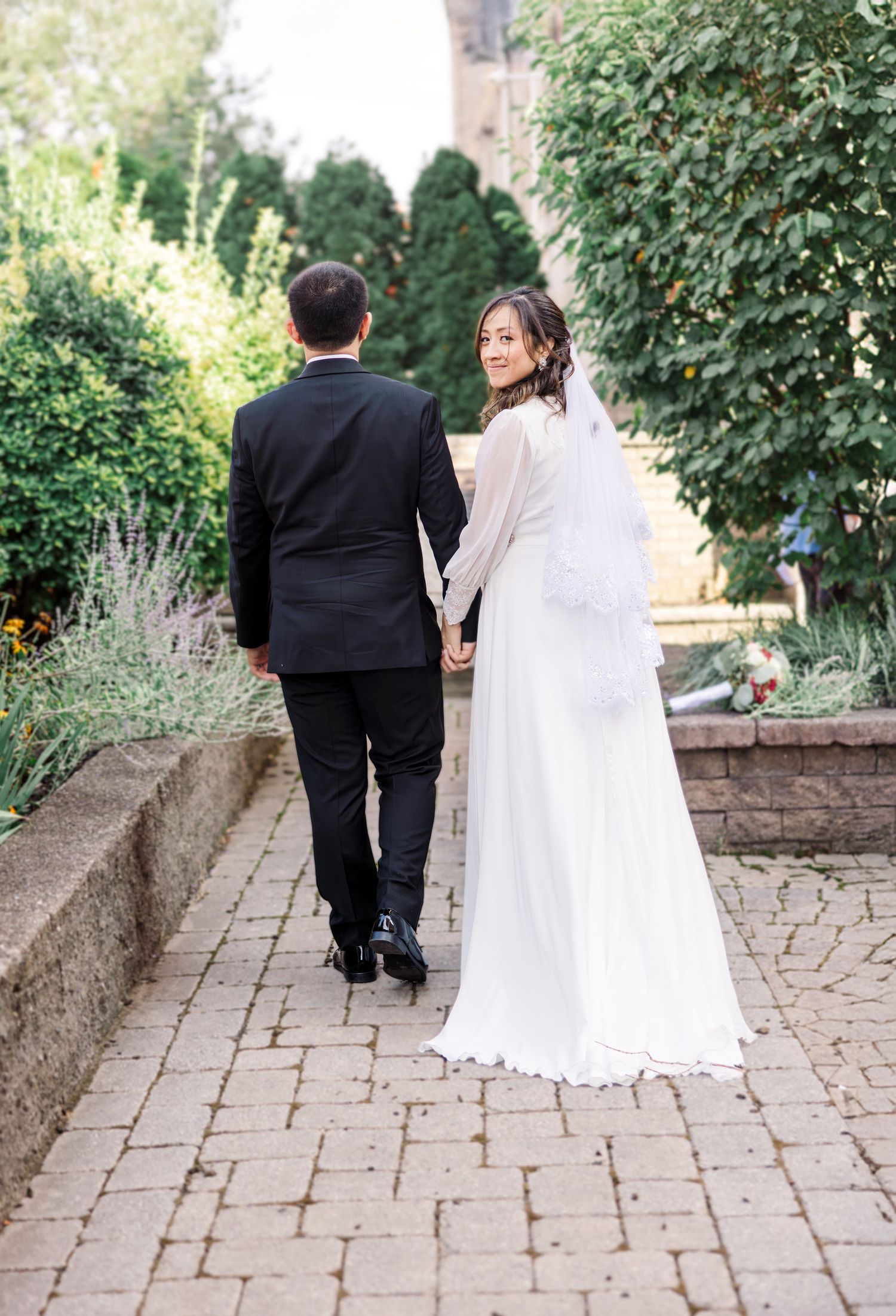 A bride in a white gown and veil holds hands with a groom in a black suit as they walk along a brick path.