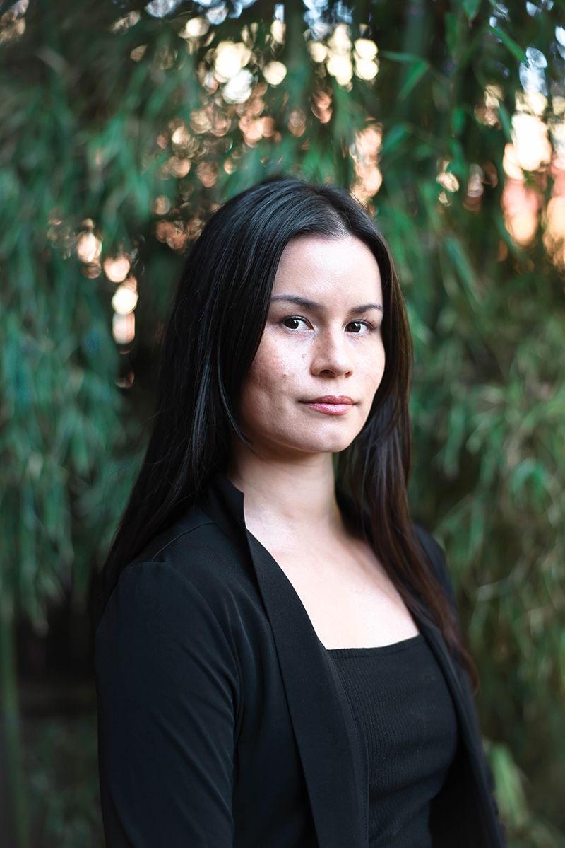 Young woman with long dark hair wearing a black outfit, posing outdoors with green foliage background.
