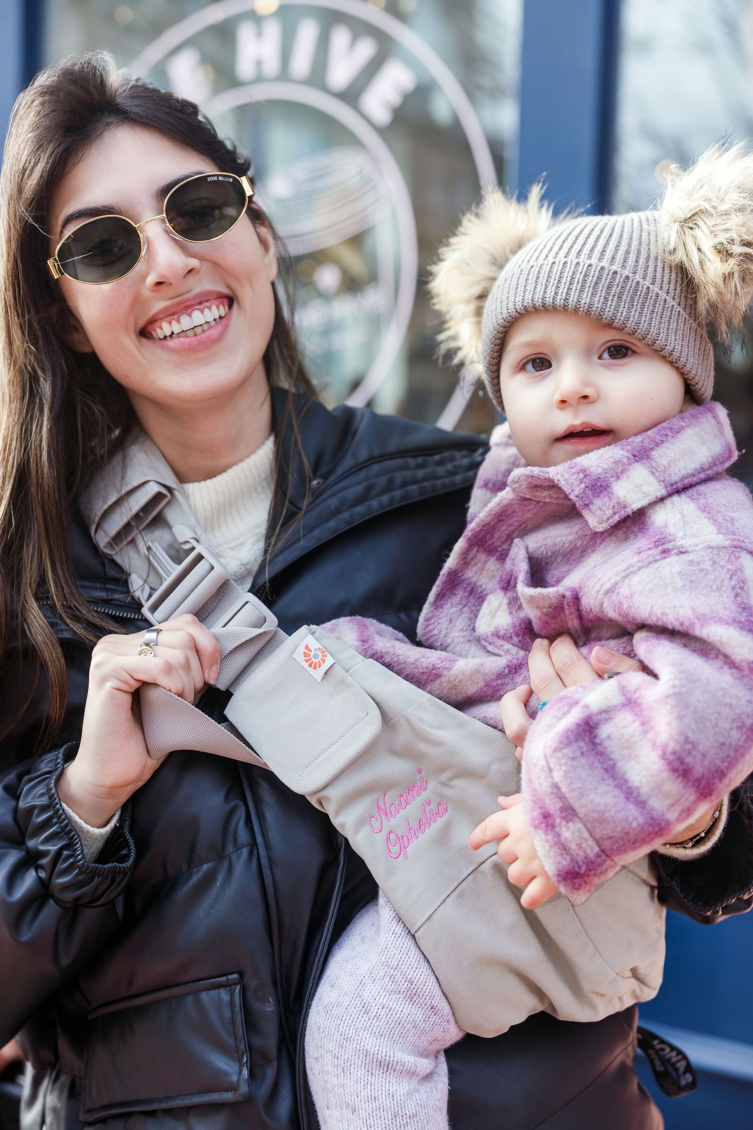 Smiling mother in sunglasses holds bundled baby in Ergobaby carrier outdoors at The Hive.