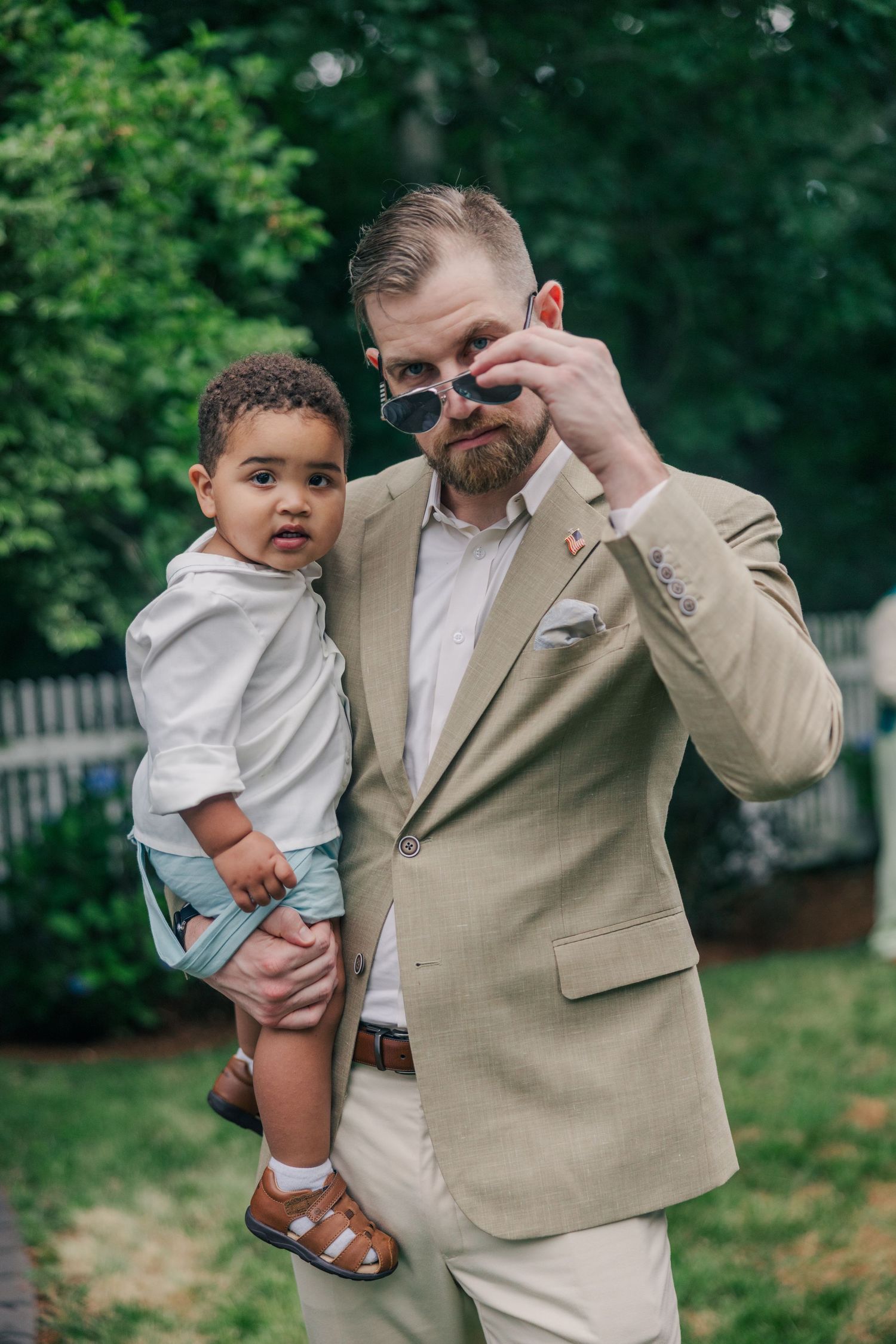 Man in beige suit holding a toddler while adjusting sunglasses outdoors in a lush green garden.