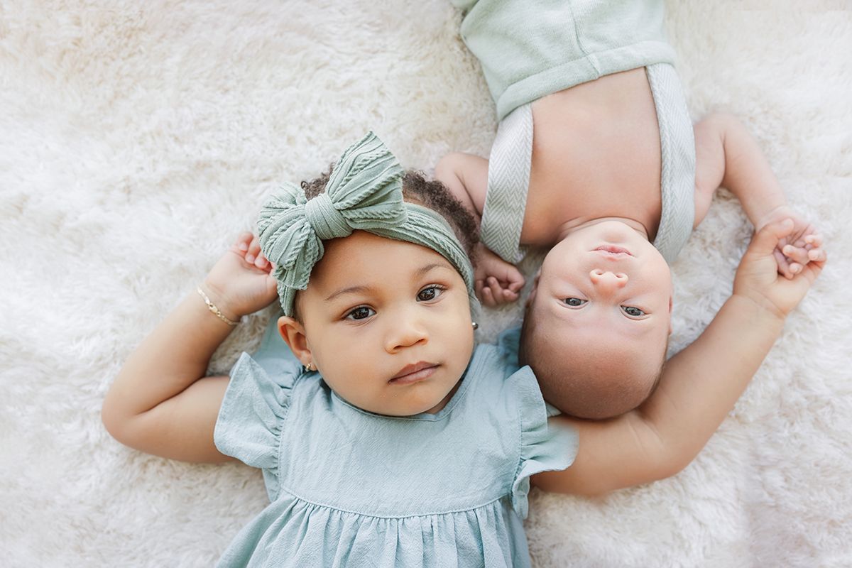 Two babies lying on a fluffy white blanket, one wearing a green bow headband and blue dress.