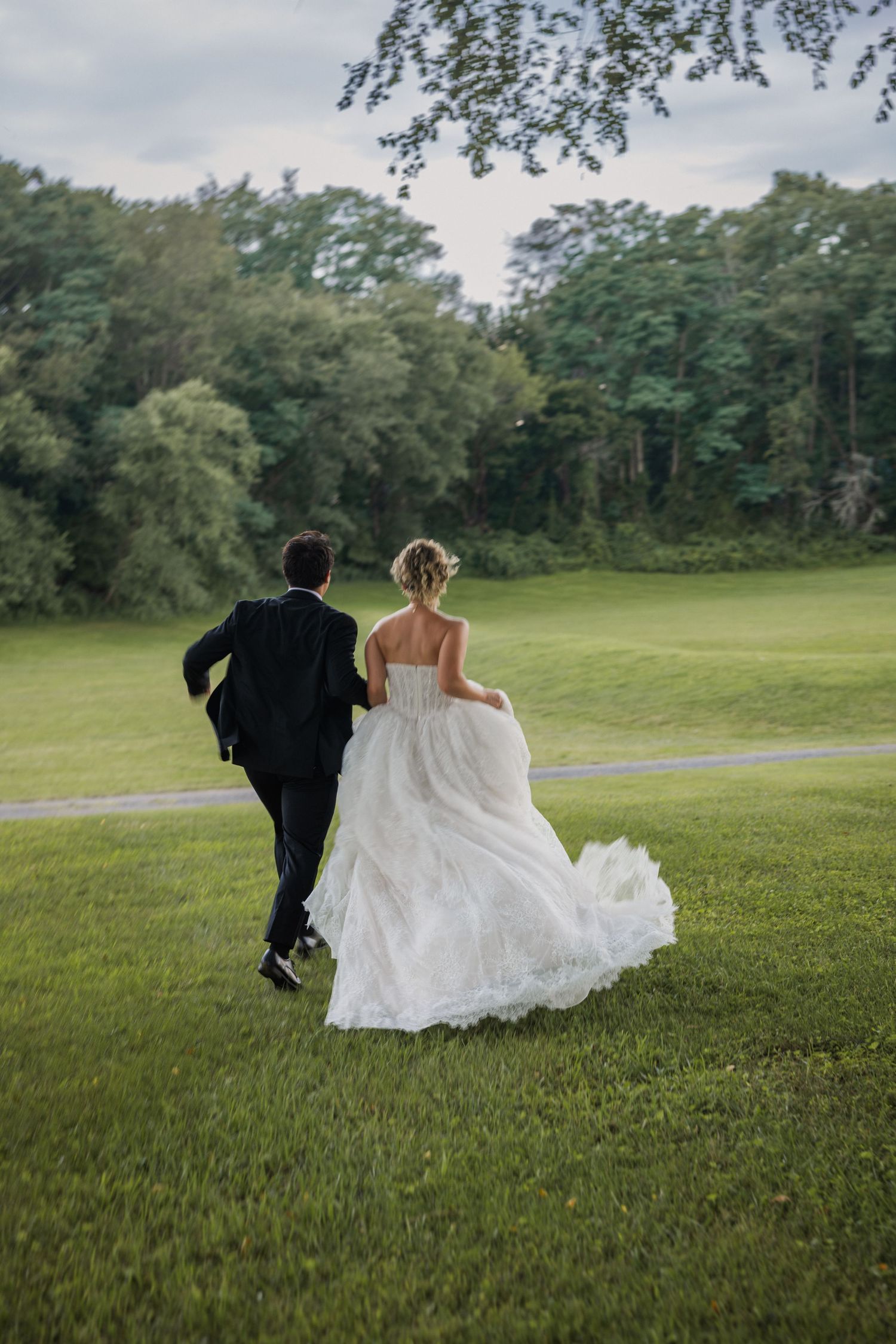 Bride and groom running across lush green lawn, bride in flowing white gown, groom in dark suit, trees in background.