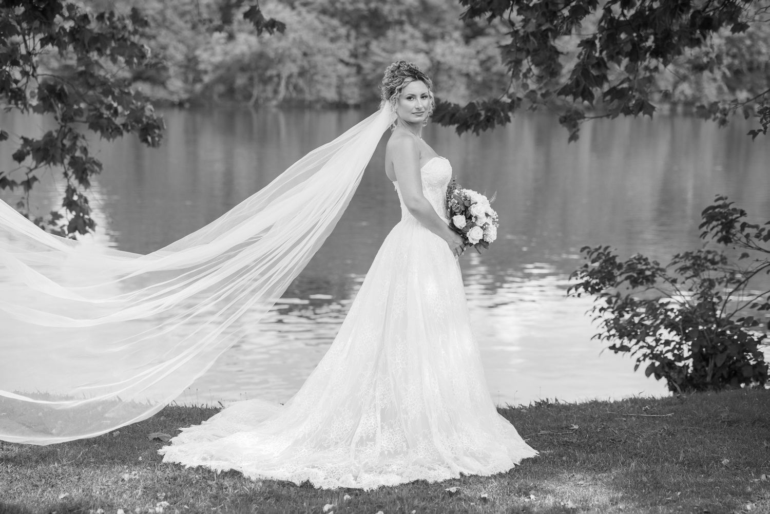 Black and white photo of a bride in a flowing gown with veil billowing, holding bouquet near a serene lake.