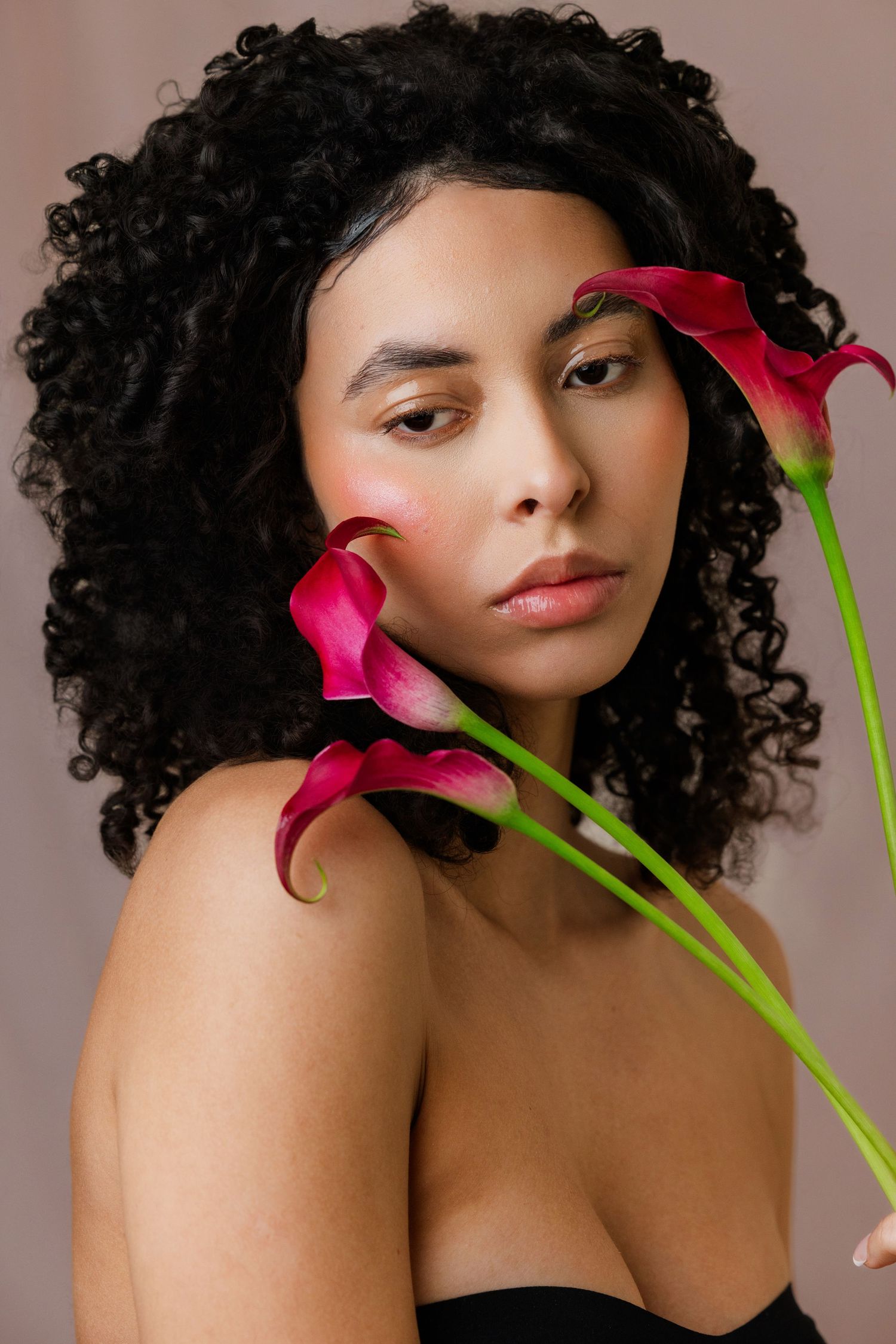 Woman with curly black hair posing with pink calla lily flowers against beige background.