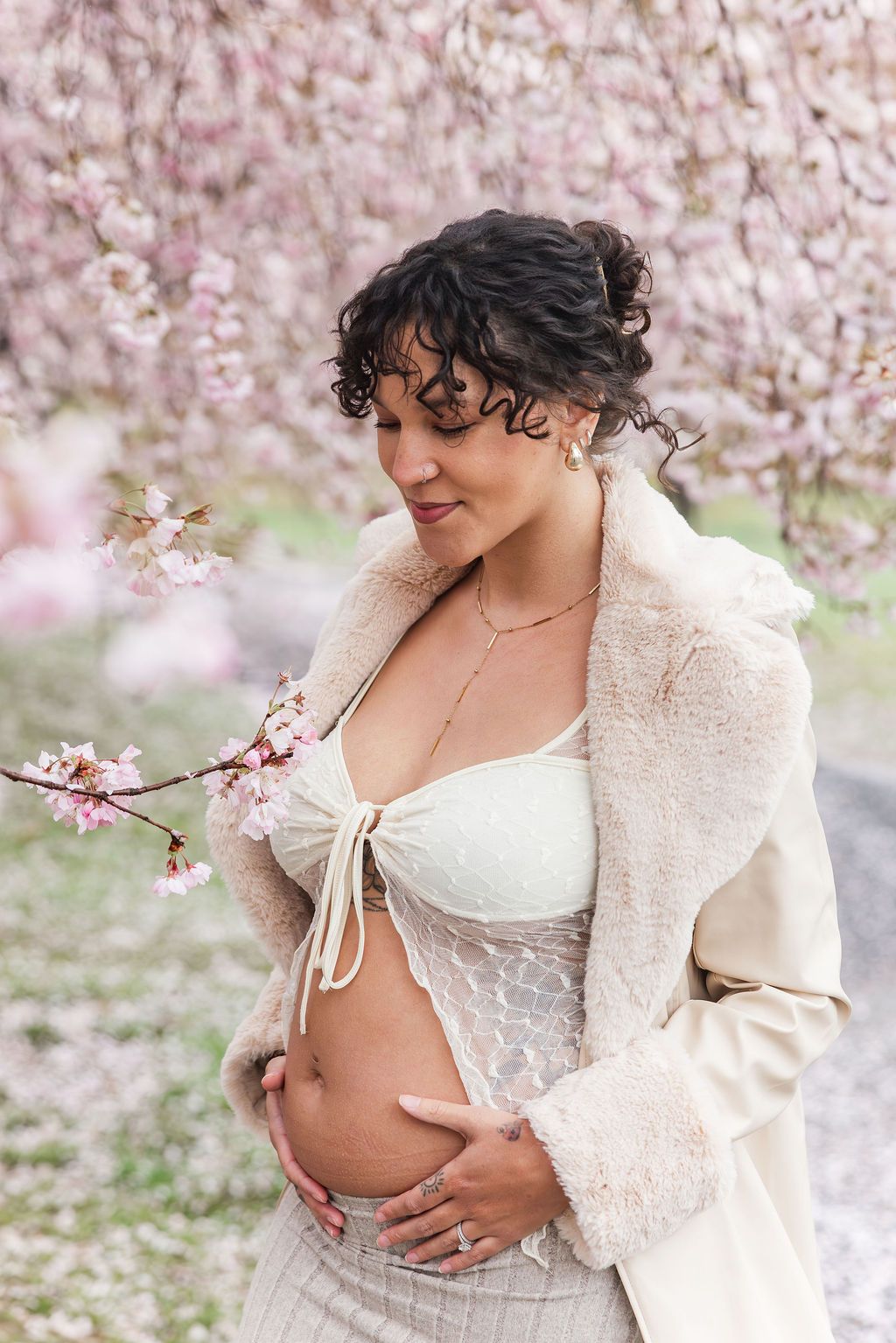 Pregnant woman in white crop top and beige coat cradles her baby bump among pink cherry blossoms.