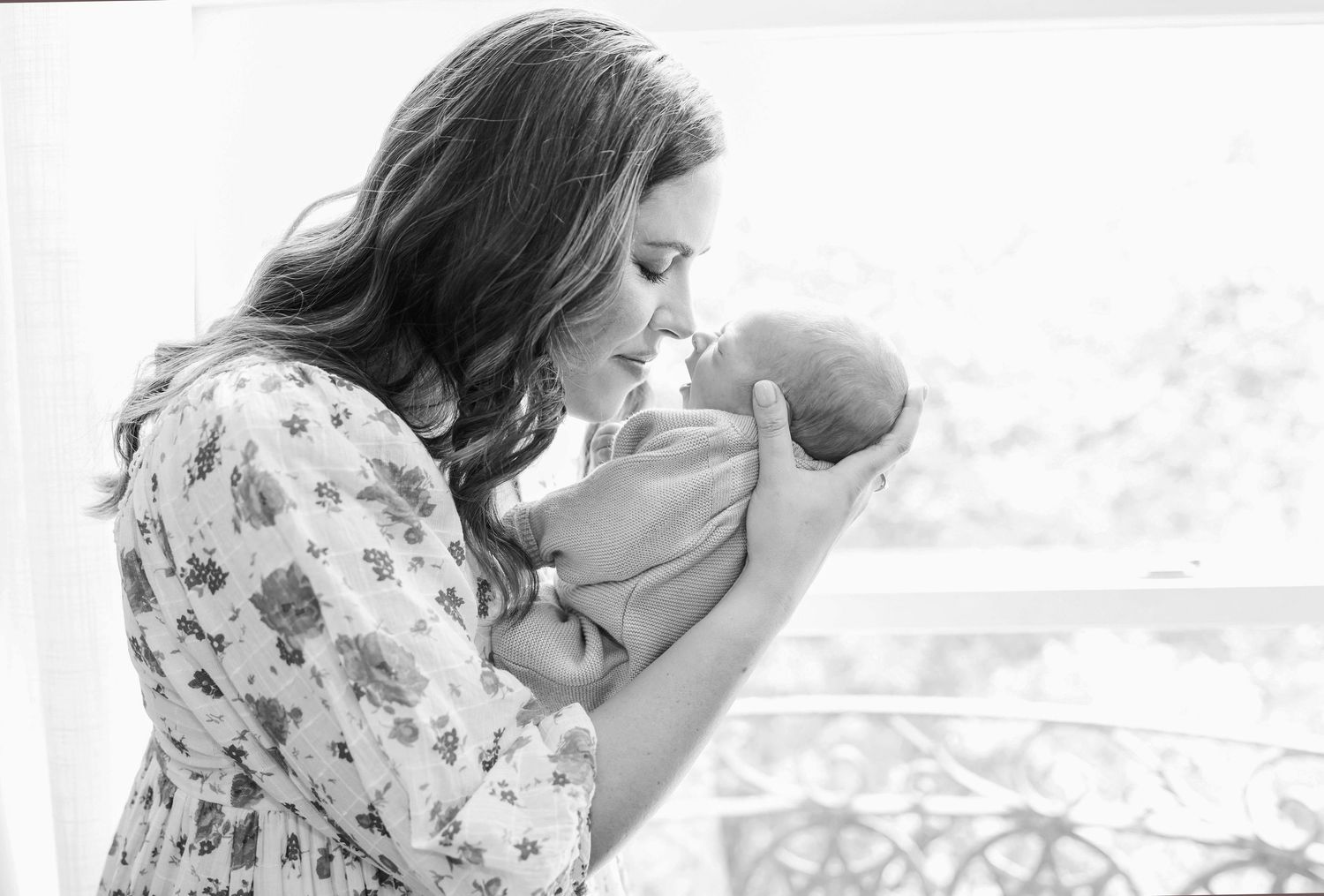 Black and white photo of a mother kissing her newborn baby near a bright window.