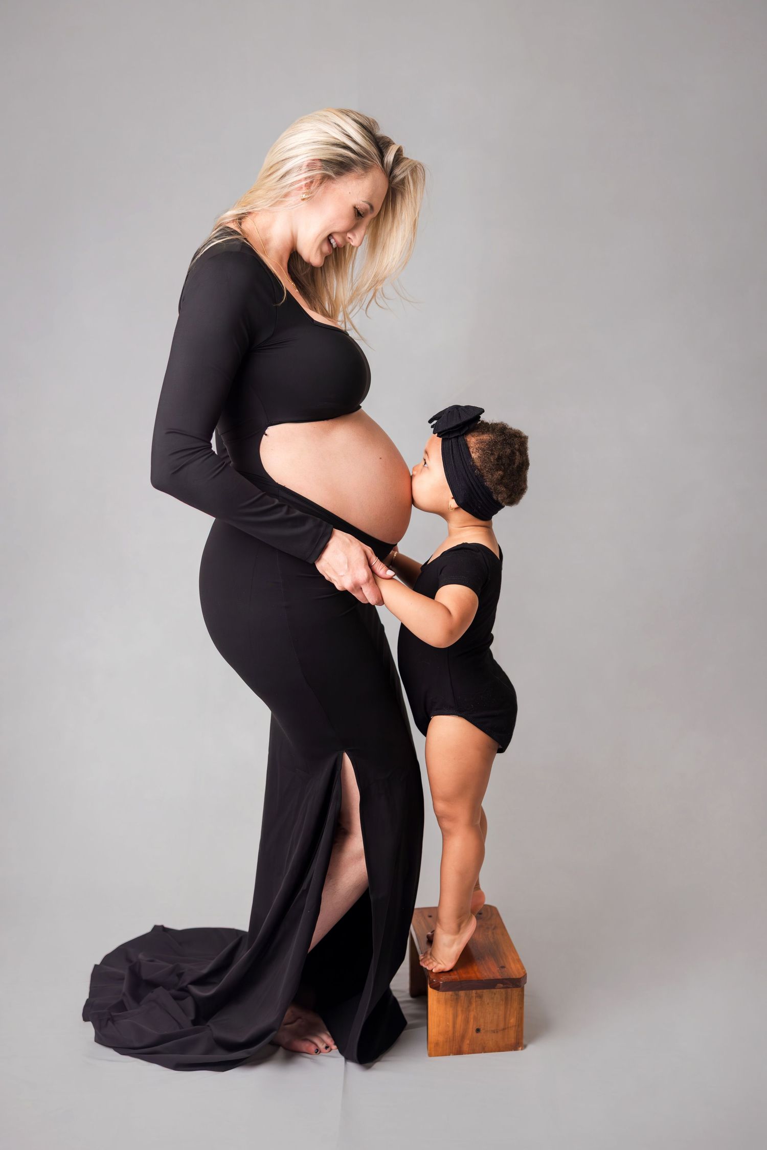 Blonde pregnant woman in black gown smiles as toddler girl kisses her bare baby bump.