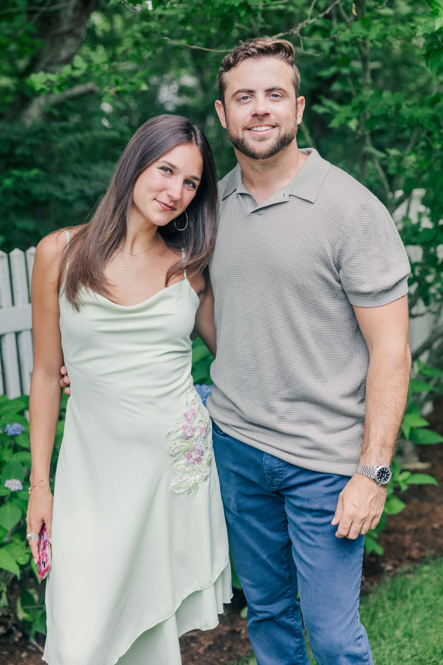A smiling couple poses together outdoors in a garden, woman in mint dress, man in gray polo and blue jeans.