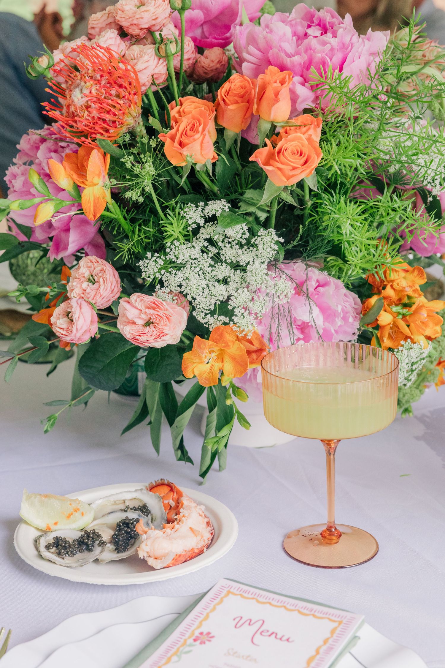 Elegant table setting with vibrant floral centerpiece, yellow cocktail, caviar appetizer, and menu card.