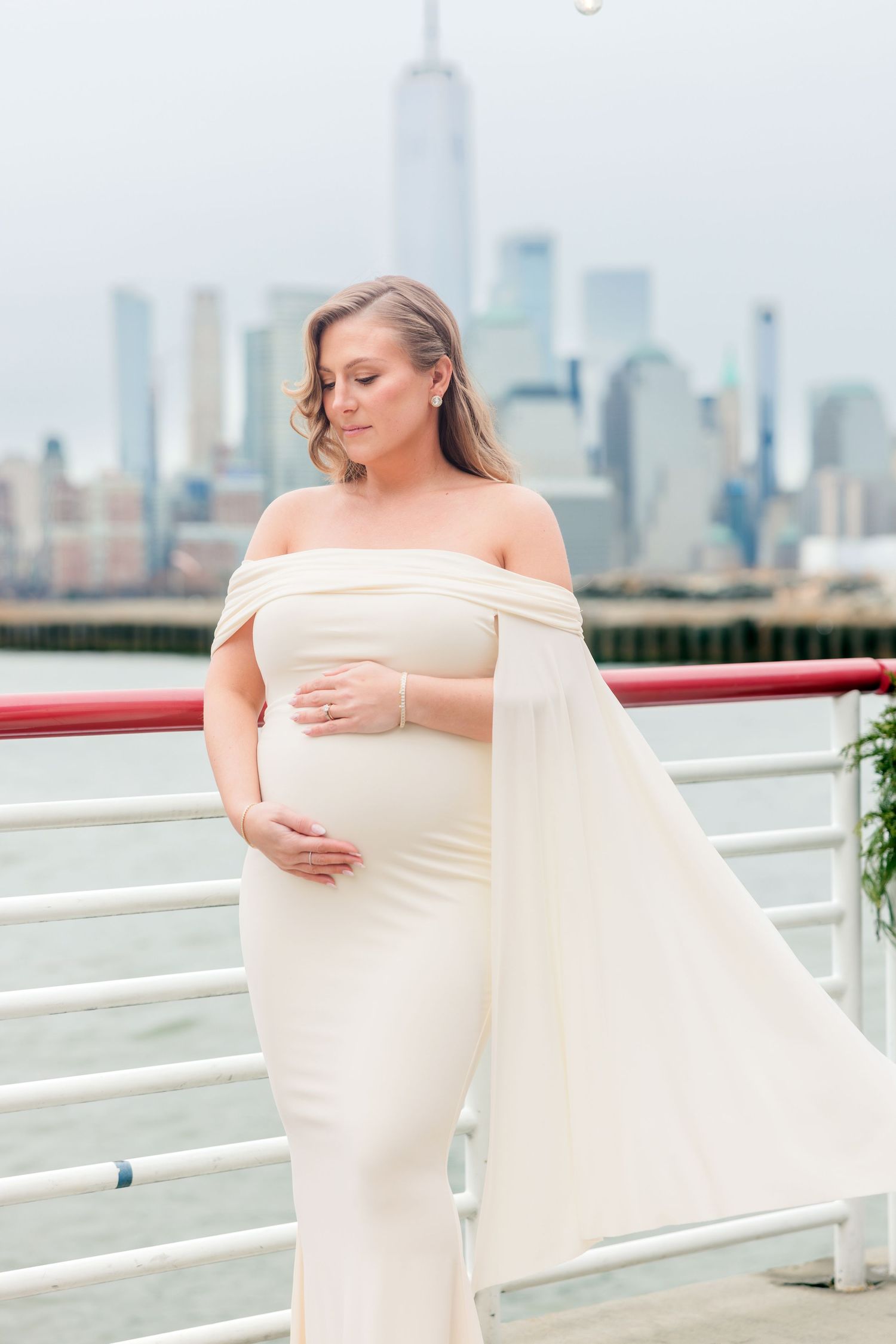 Elegant pregnant woman in white off-shoulder gown holds baby bump with NYC skyline in background.