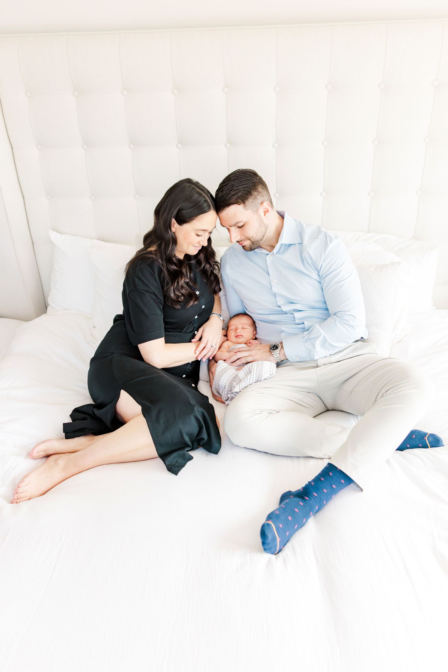 New parents sitting on white bed smiling at their newborn baby wrapped in pink swaddle.