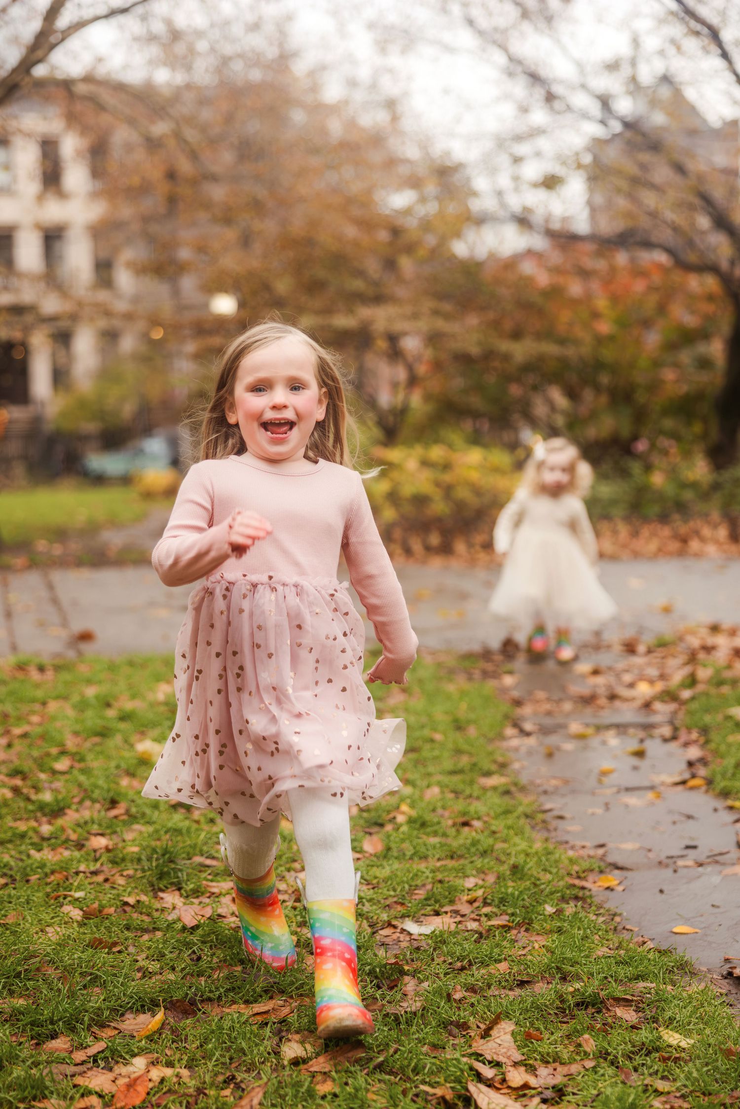Joyful little girl in pink tutu and rainbow boots running through autumn park with sister behind her.