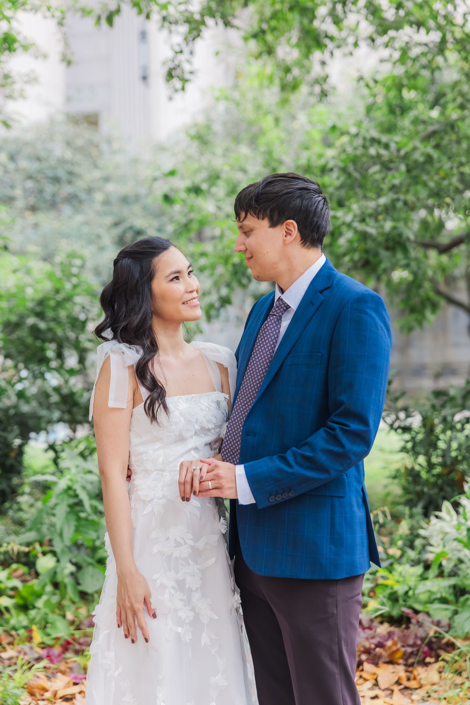 Asian bride and groom in blue suit share a tender moment holding hands in a garden setting.