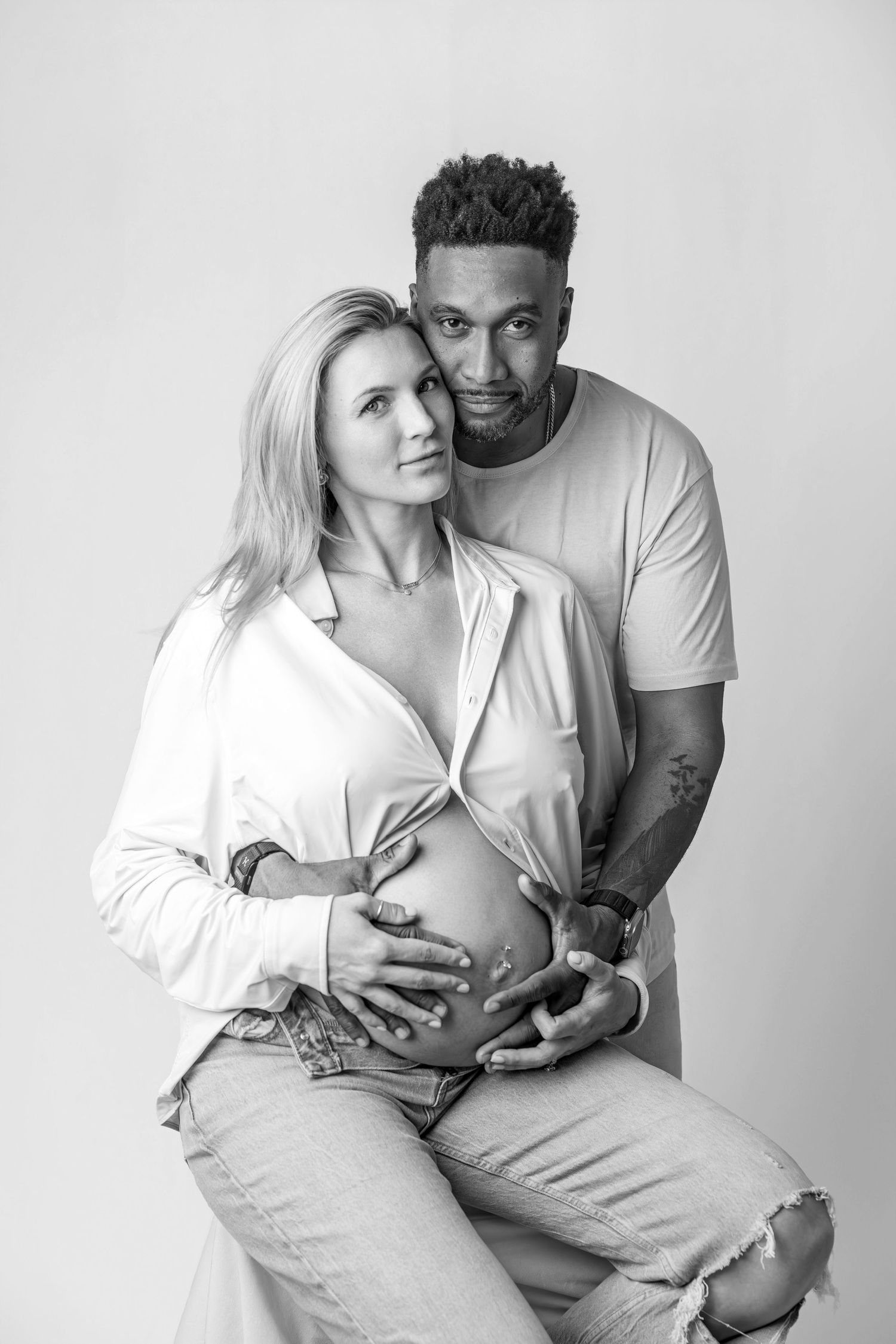 Black and white maternity portrait of a couple gently holding a pregnant belly in a studio setting.